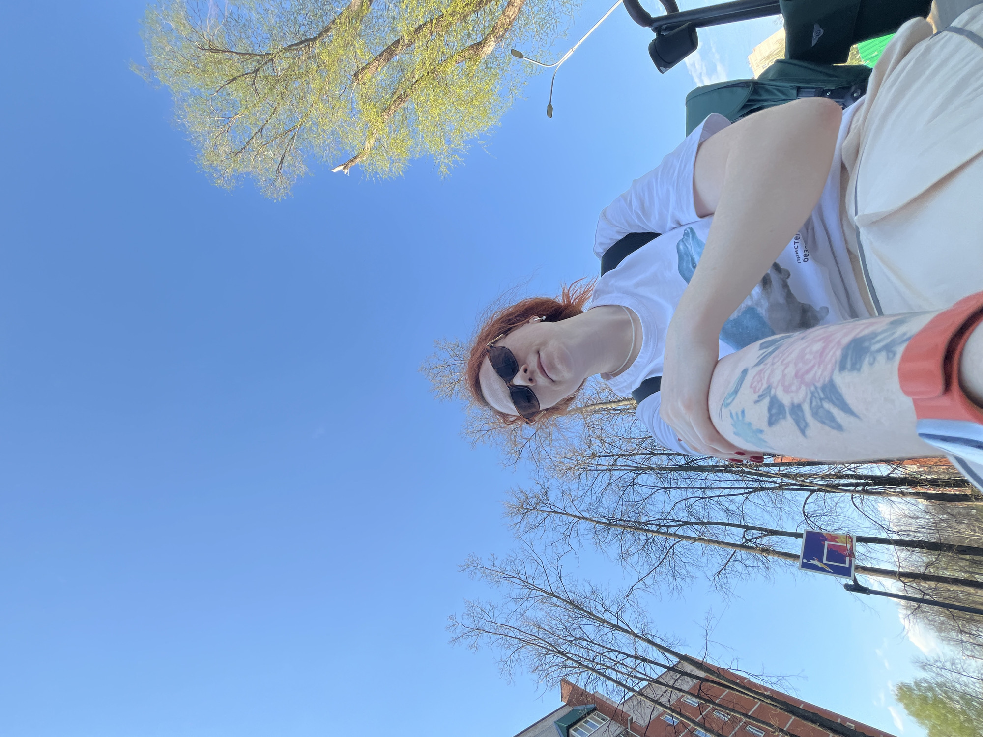 The image is taken from a low angle looking upward. A person with red hair and sunglasses is seated and facing the camera, wearing a white shirt and tan shorts. The person has a tattoo on their arm. Above, there's a clear blue sky partially obscured by tree branches, some with green leaves and others bare. In the background, there is a building made of brick and concrete and a street sign that indicates a pedestrian crossing. The photograph appears to have been taken in a park or urban green space. The location is possibly in Chaikovsky, Russia.