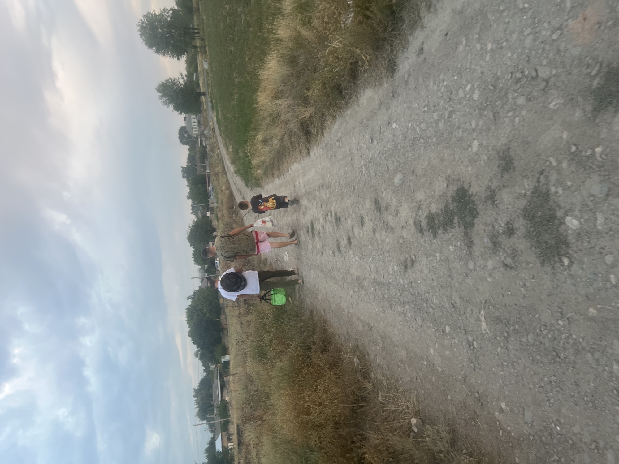 Three individuals, two adults and one child, are walking away from the camera down a gravel road. The adult on the left wears a white t-shirt and dark pants and carries a green bucket and a backpack. The adult in the middle wears a camouflage t-shirt and pink and white patterned shorts. The child on the right wears a black t-shirt with a cartoon character and dark shorts. The road is bordered by dry grass and vegetation on the left and a green grassy field on the right. In the background, there are houses and trees, suggesting a rural or semi-rural setting. The sky is overcast. The location is identified as Дачное, Kyrgyzstan.