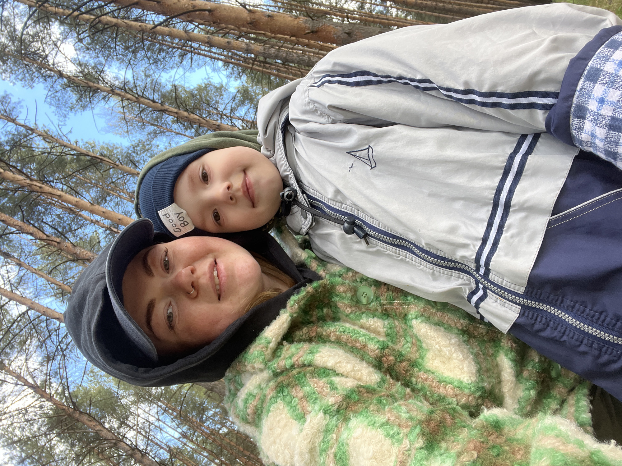 A low-angle selfie shows a woman wearing a black hooded sweatshirt and a green and cream patterned jacket, and a young boy wearing a blue hat labeled "Good Boy" and a light gray jacket with a sailboat emblem. The background consists of tall trees and a blue sky with white clouds. The location is most likely Chaikovsky, Russia, based on the prompt.