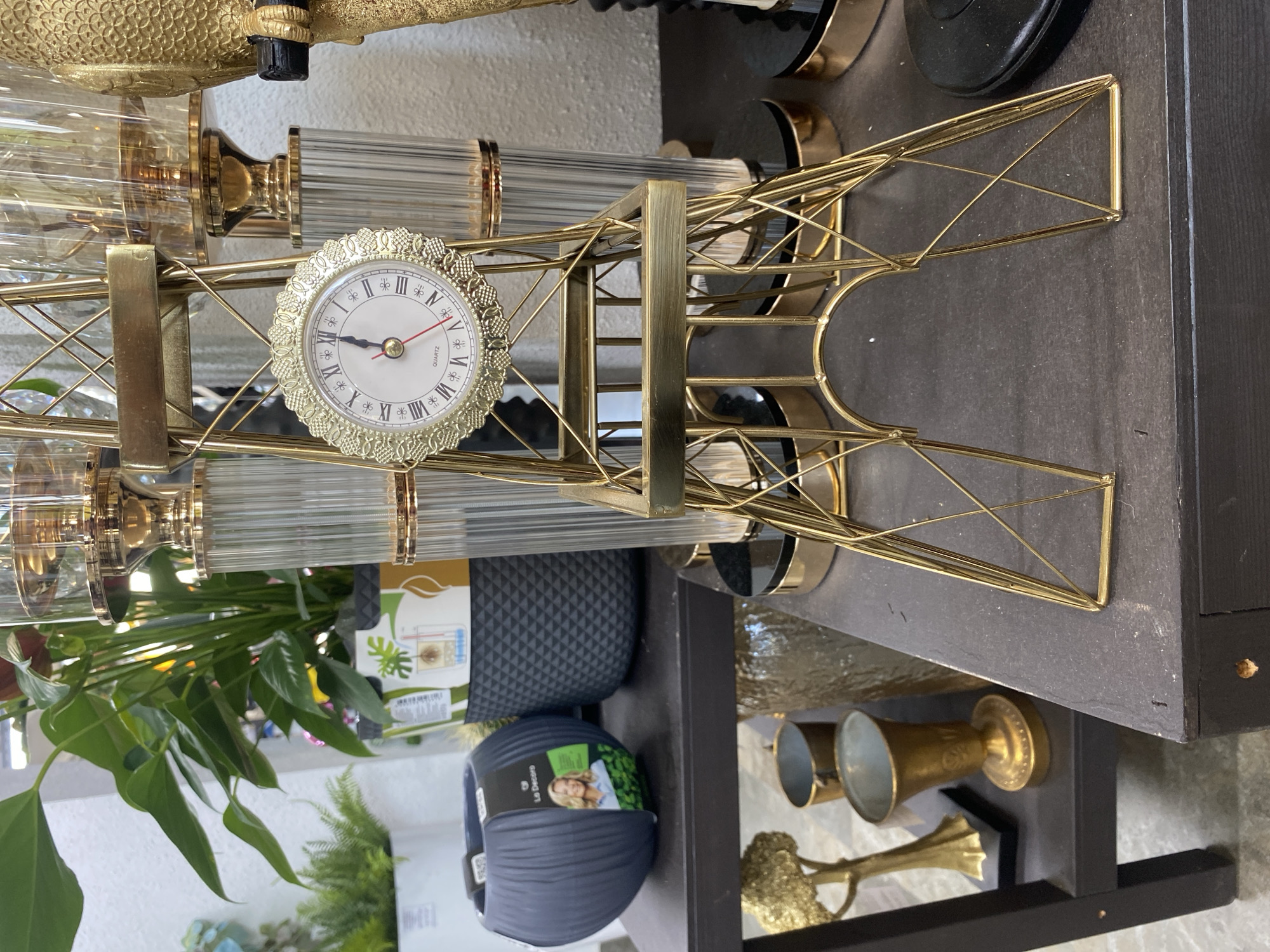 The image shows a golden Eiffel Tower replica with a clock face. It is placed on a dark surface, potentially a shelf or display stand. The setting is indoors, likely a shop or home decor store. Additional details include decorative glass elements, plant pots, and other golden objects in the background, creating a rich, ornate aesthetic. While a precise location within Yekaterinburg, Russia, cannot be determined from the image alone, the style of decor suggests a store or residence with a taste for classic, European-inspired design.