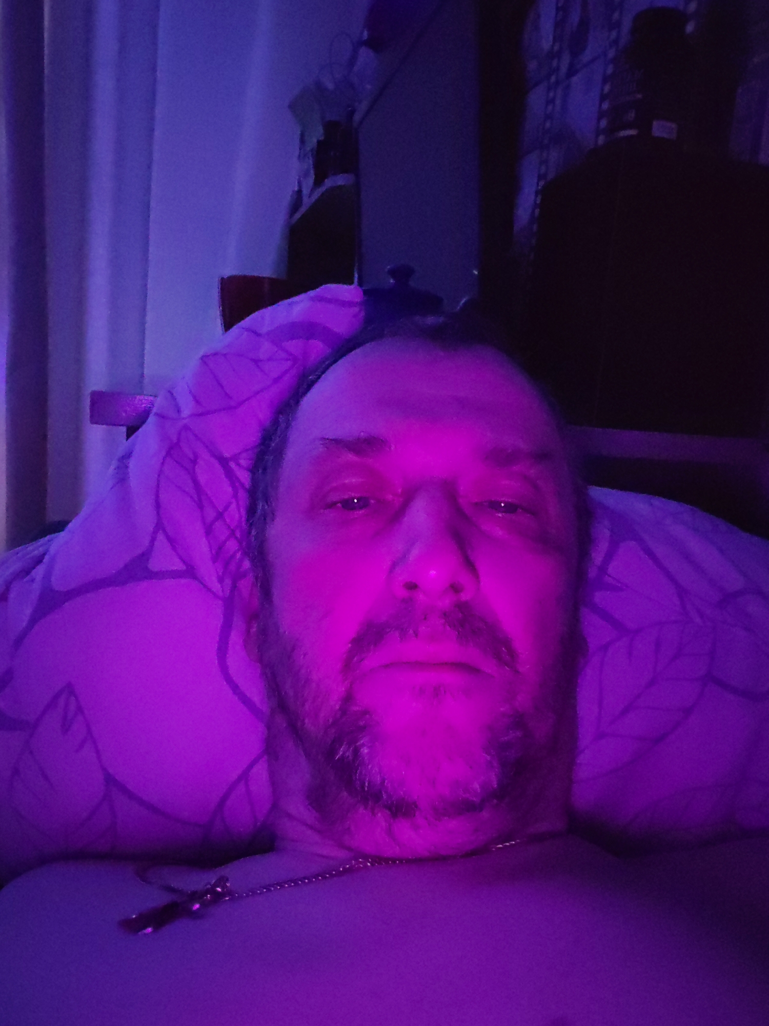 A medium close-up photograph shows a middle-aged man lying in bed with his head on a leaf-patterned pillow. He has a beard and wears a necklace with a cross pendant. The lighting is predominantly purple, which obscures details in the background. In the background, there is a partially visible curtain, some furniture, and an indiscernible object against what appears to be patterned wallpaper. The location of Chaikovsky, Russia, is not discernible from the image.