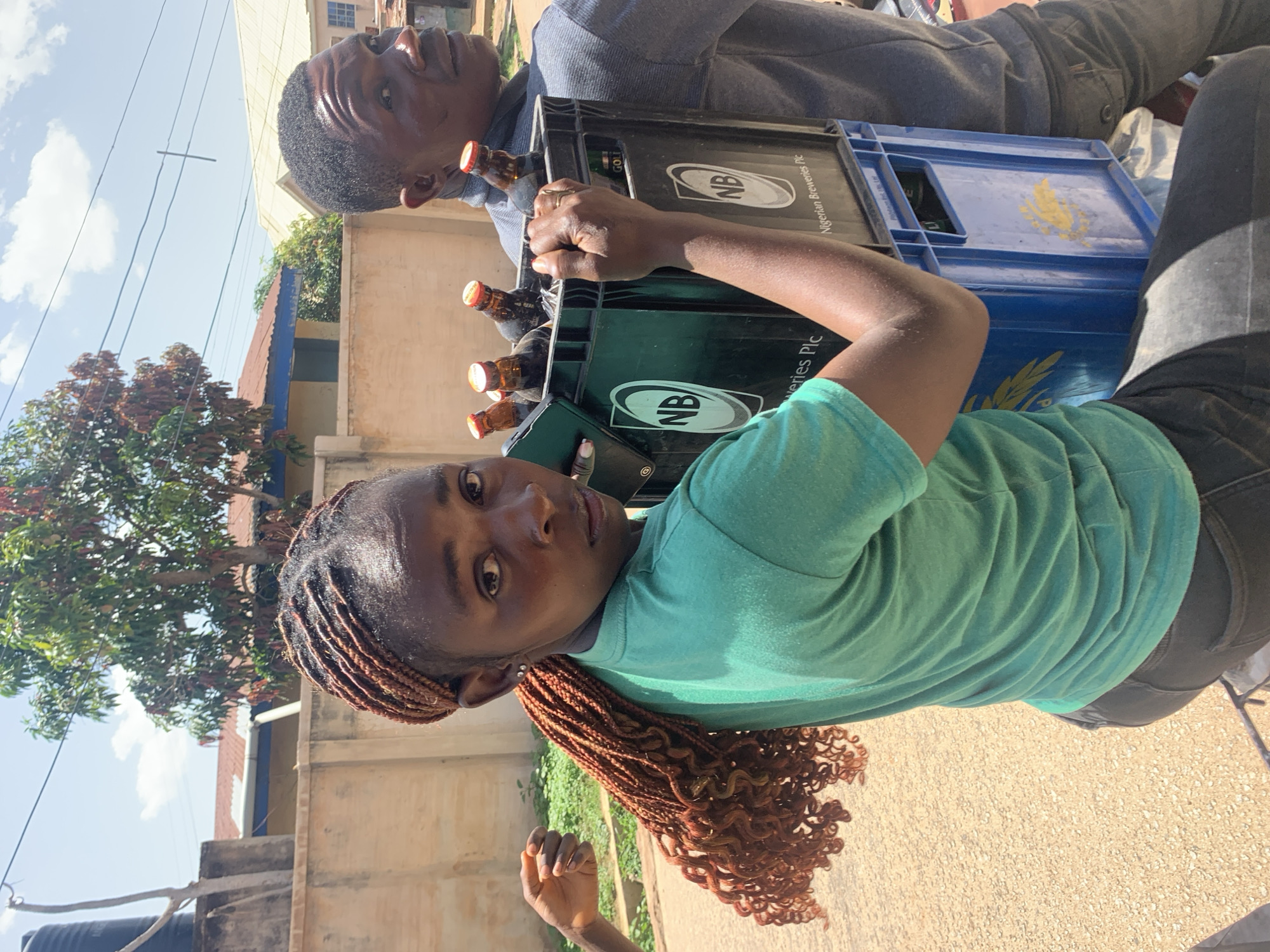 In Jos, Nigeria, two individuals are seen outdoors. A young woman with braided hair in a turquoise t-shirt and dark pants carries a black crate containing several bottles of beer, with a blue crate also visible. She is holding the black crate with her right hand. Beside her, a man in a gray shirt and dark pants walks along, partially obscured. In the background, a beige wall with some vegetation and buildings can be observed, along with a water tank and power lines. The ground is covered in sand and gravel.