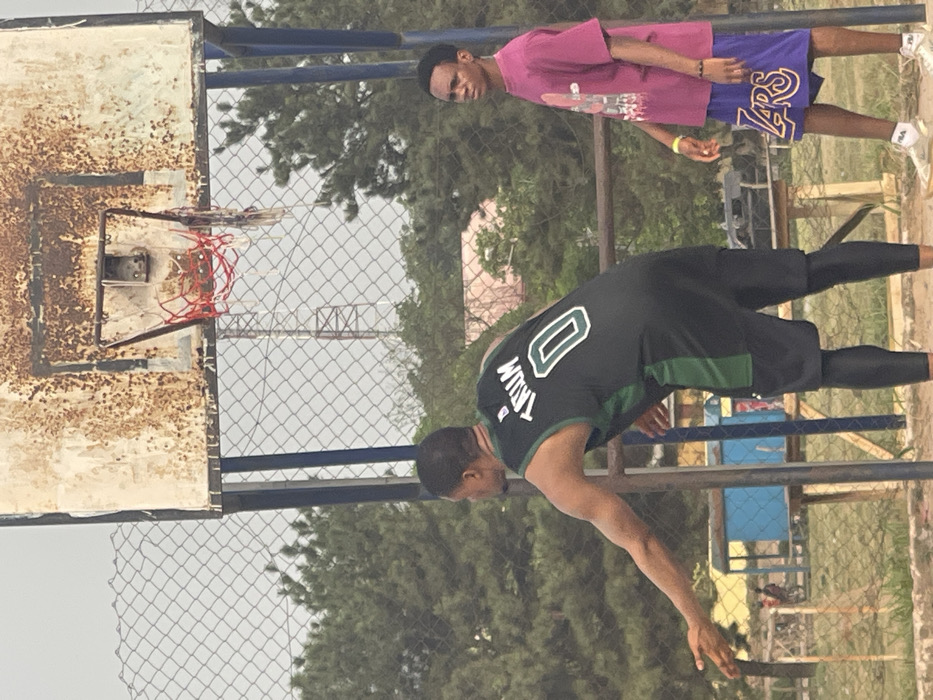 The image depicts an outdoor basketball court in Umunekwu, Nigeria, under an overcast sky. In the foreground, two adult males are visible. The individual on the left, viewed from the back, wears a dark green/black basketball jersey with "TRUM" and the number "0" in white, black shorts, and black compression tights. His right arm is extended downwards and slightly outwards, hand open. To the right, a second individual stands facing left, wearing a magenta t-shirt with a white graphic, purple shorts with "LAKERS" in gold lettering, and light-colored sneakers. A yellow wristband is present on his right wrist.
The basketball equipment includes a heavily rusted, peeling white backboard with a red rim and a partially torn red and white net. A dark blue support pole for the backboard is visible. Behind the court, a chain-link fence separates the playing area from the background. Visible through the fence are green trees, a distant metal lattice tower, a small structure with a blue base, and a light pink/orange building. The ground surface appears to be dirt or earth.