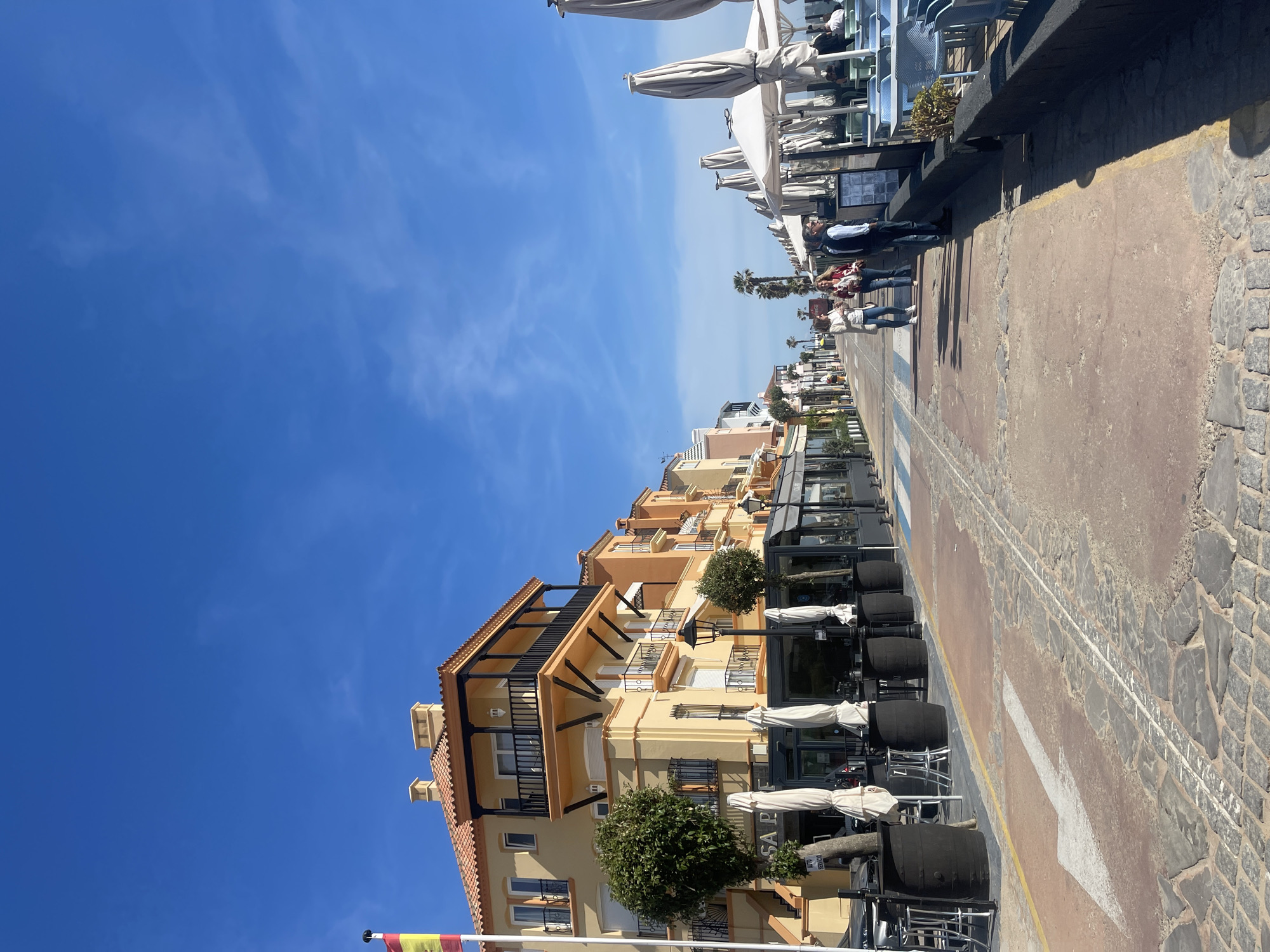 The image depicts a street scene in El Puerto de Santa María, Spain. On the left side of the frame are buildings with a yellow and orange facade, adorned with balconies and topped with red tiled roofs. Restaurant seating and umbrellas are visible along the street, with planters adding greenery.
On the right side of the image is a sidewalk and a street, marked with a white arrow. A few people are walking along the sidewalk. The sky is clear and blue. The overall impression is of a sunny day in a coastal city.