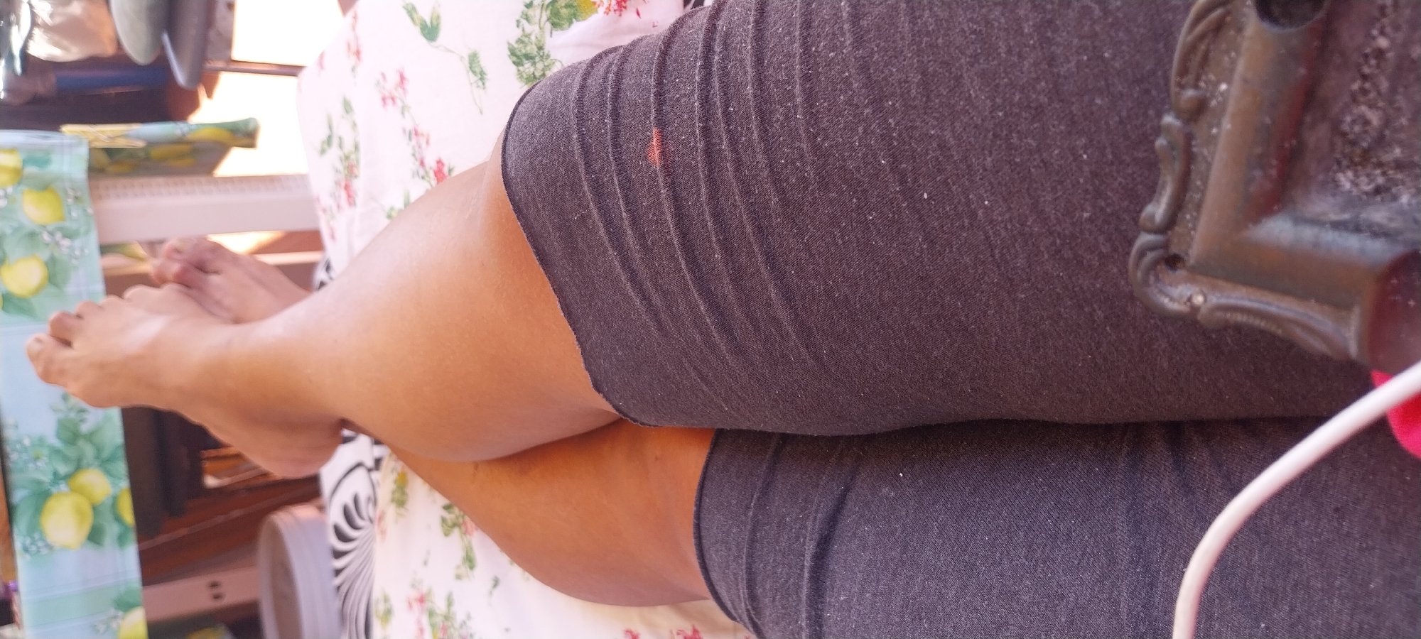 The image shows a close-up of a person's legs and feet, which are crossed. They are wearing dark gray leggings or shorts. The legs are resting on a surface covered with a white and floral patterned tablecloth. In the background, a table covered with a tablecloth featuring lemons and green leaves is visible. The setting appears to be outdoors, possibly a patio or garden, given the presence of natural light and the casual setting. It is impossible to determine the specific location in Ardea, Italy, from the visual information provided.