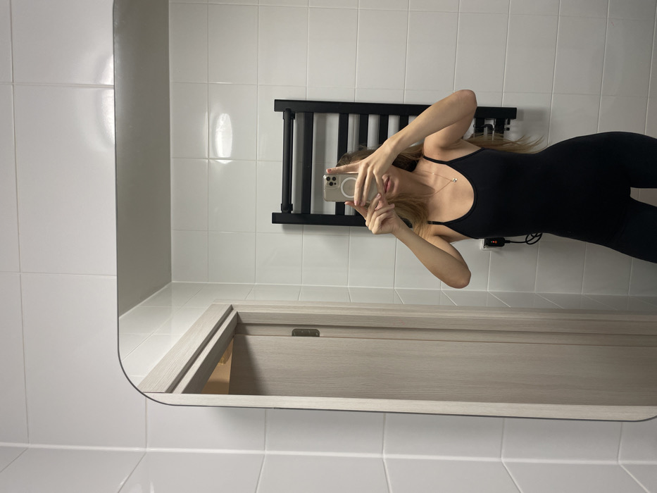 A mirror shows a woman in a black jumpsuit holding a phone in front of her face, presumably taking a photo. The background consists of white tiled walls and a dark grey towel rack. A doorway is visible below the woman's reflection, featuring a light-colored, closed door. The overall setting appears to be a bathroom.