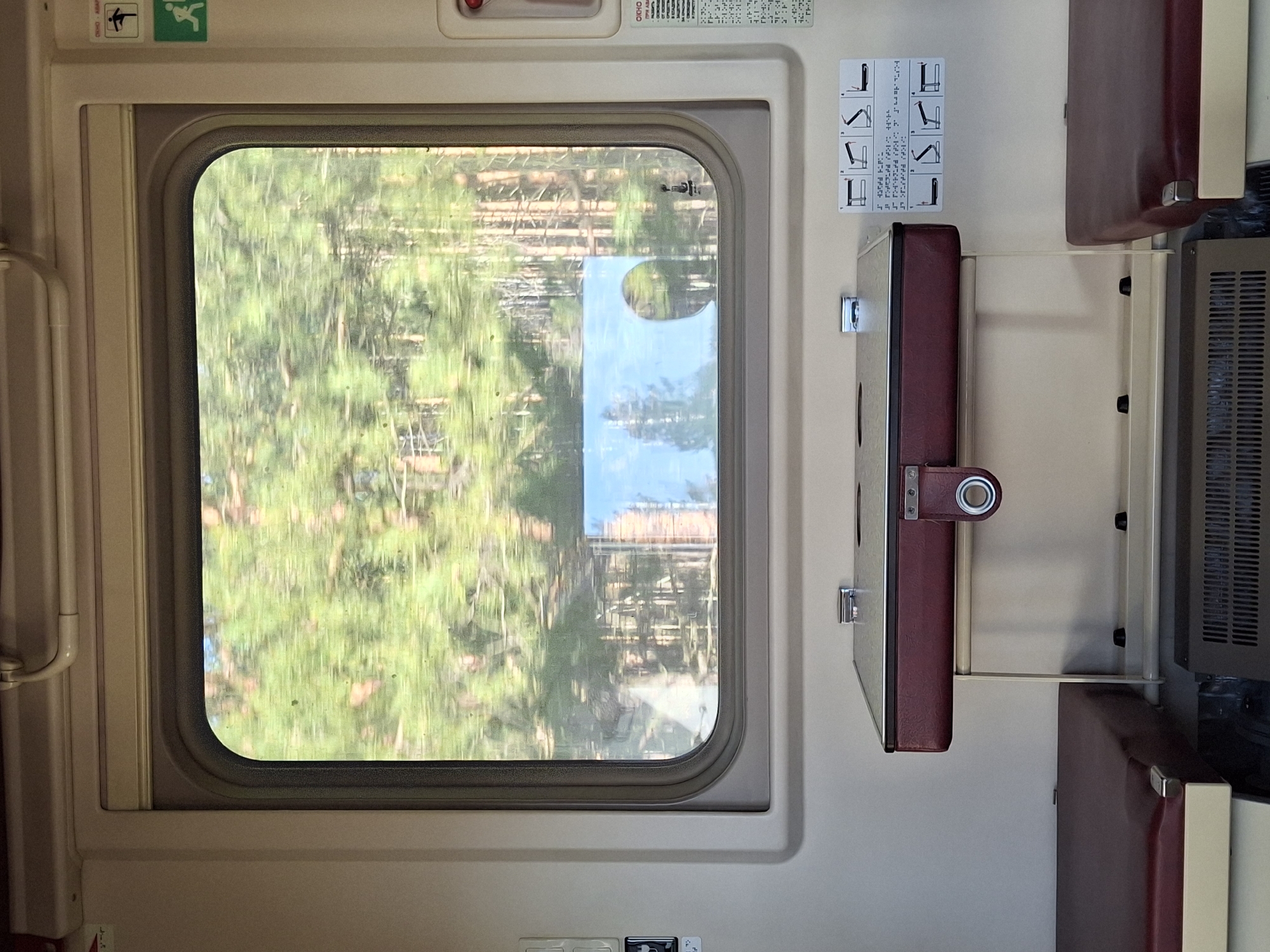 Here is a forensic description of the image:
The image shows the interior of a train car, likely in motion, given the blurred landscape seen through the window. The focus is on the window, with trees and other vegetation passing by rapidly outside. A small, folded-down table is attached to the wall next to the window. A set of instructions, potentially related to the table or seat configuration, is posted on the wall above the table. There is a radiator vent to the right. The walls are pale and utilitarian. The seats are upholstered in a dark reddish-brown material, with cream-colored trim. The scene is devoid of people, suggesting the photo was taken in an empty train car or section. No specific location can be determined from the image.
