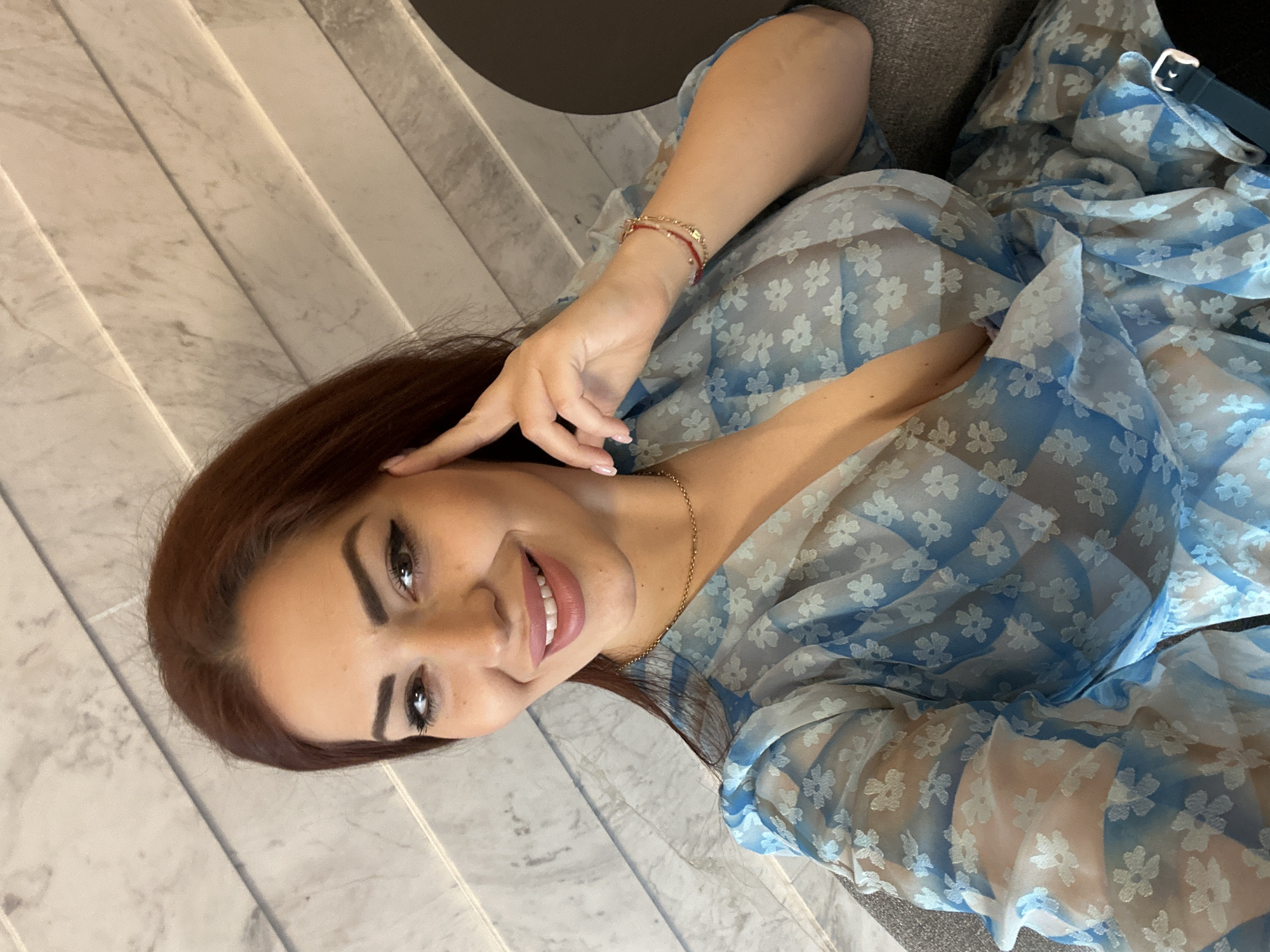 A woman with auburn hair is smiling at the camera. She is wearing a blue and white floral print dress. She is wearing a thin gold necklace and two thin bracelets, one gold and one red, on her right wrist. She has dark, arched eyebrows and defined eyelashes. Her right hand is raised near her temple. She appears to be sitting indoors on a gray fabric surface with a patterned wall in the background. The location is Kazanlak, Bulgaria.