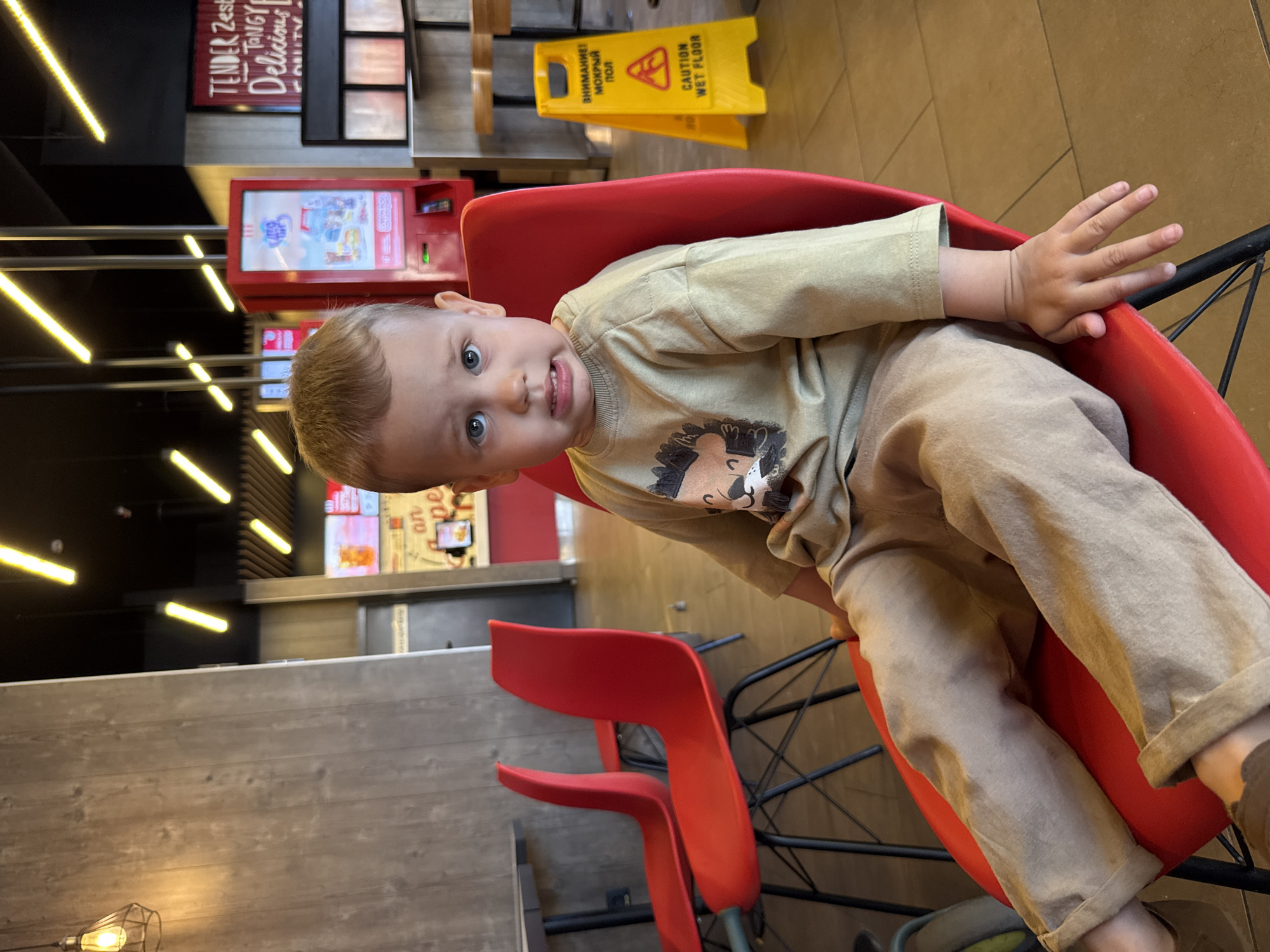 This image depicts a young child seated in a red chair within a fast-food restaurant located in Rusanovo, Russia. The child, a toddler with short light-colored hair and blue eyes, looks directly at the camera with a slightly pensive or uncomfortable expression, exhibiting slightly parted lips and a furrowed brow. They are wearing a light olive-green long-sleeved shirt with a dark, stylized animal graphic on the front and loose-fitting khaki-colored pants.
The restaurant interior features a dark ceiling with numerous diagonal linear yellow light fixtures. To the left, a faux-wood paneled wall and a pendant light are visible. In the background, other red chairs and dark tables are present. On the right, a yellow A-frame 'CAUTION WET FLOOR' sign is prominent, displaying both English and Russian text ('ВНИМАНИЕ! МОКРЫЙ ПОЛ'). Behind it, a red self-order kiosk displaying a colorful advertisement is visible, alongside a large red sign with white text, partially reading 'TENDER Zest Tangy Delicious'. The floor is composed of light-colored tiles.