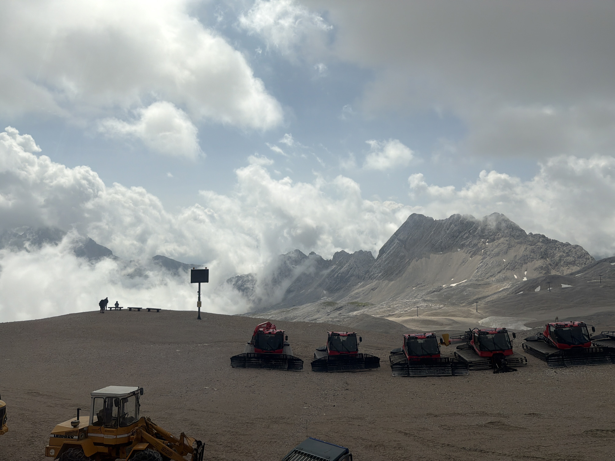 The image depicts a high-altitude, mountainous landscape near Garmisch-Partenkirchen, Germany. The foreground features a sandy or gravelly terrain with several snow groomers (also known as piste bashers) parked in a line. These are large, red vehicles with tracks, designed for maintaining snow surfaces. To the left, a yellow bulldozer-like vehicle with the brand name "LIEBHERR" is partially visible. In the middle ground, a small group of people are standing and sitting on benches on a slight incline, with a signpost nearby. The background is dominated by a rugged mountain range, partially obscured by dramatic cloud formations. The sky is a mix of blue and heavy, grey clouds. The overall scene suggests a ski resort or a mountain area at a significant elevation, possibly during the off-season or a cloudy day.