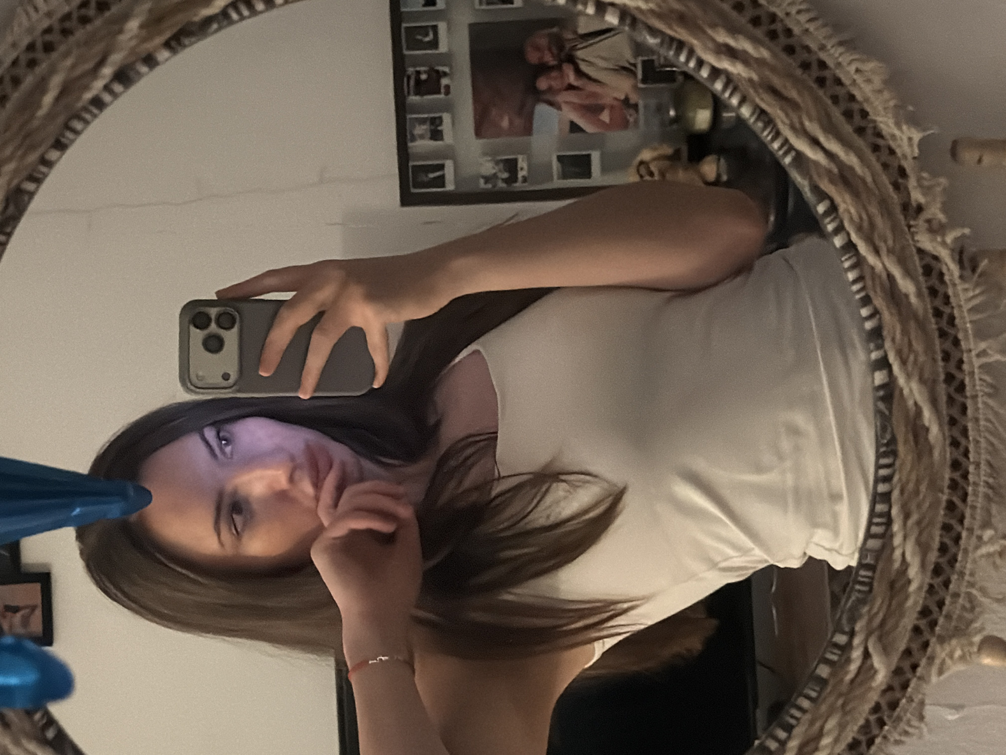 A woman with long brown hair is taking a mirror selfie indoors. She is wearing a white tank top and a red bracelet on her left wrist. Her right hand is holding a grey smartphone, and her left hand is touching her chin. The mirror has a woven, decorative frame. In the background, visible in the mirror, is a wall with framed photographs and some decorative items. There is no indication of the location being Sharm El Sheikh, Egypt.