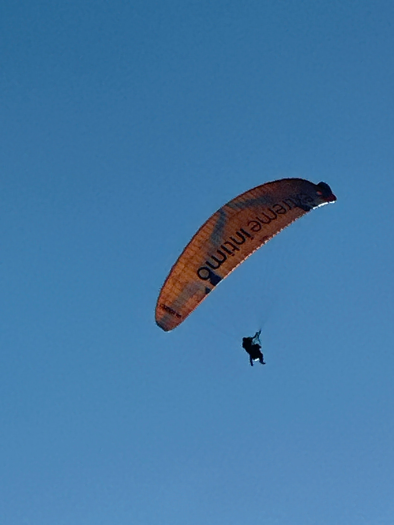 In the clear blue sky above Boreti, Montenegro, a paraglider is captured mid-flight. The paraglider's orange canopy, emblazoned with the text "extreme intima", is prominently displayed against the bright sky. Below the canopy, a solitary figure, the paraglider, is suspended, seemingly suspended by the wind. The scene is devoid of any other human activity or structures, emphasizing the vastness of the sky and the solitude of the paraglider's journey. The image captures a moment of aerial freedom against a backdrop of a serene, cloudless sky.