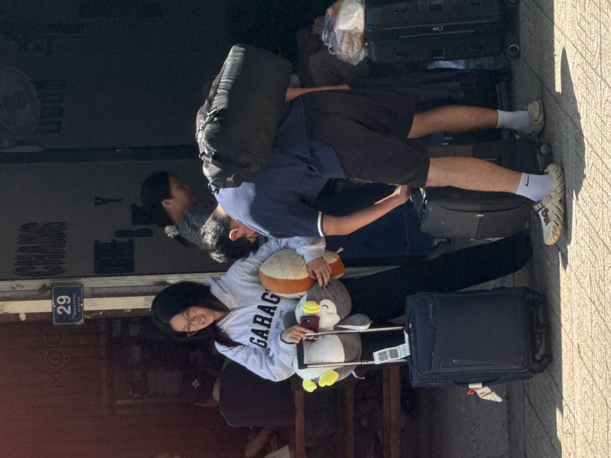 The image depicts an outdoor scene with several people and luggage, possibly at a transportation hub or a hotel entrance. In the foreground, a young woman with glasses, wearing a grey hoodie and black pants, is holding a stuffed animal and a luggage handle. Next to her, a person in a navy blue polo shirt and black shorts is bending over to adjust luggage, with a large black duffel bag on their shoulder. Behind them, other individuals are partially visible. The environment includes a tiled ground and a building with signage that reads "MỘC 29" and "CHÀNG." The overall lighting suggests it is daytime, with shadows cast on the ground. The presence of multiple suitcases indicates travel.