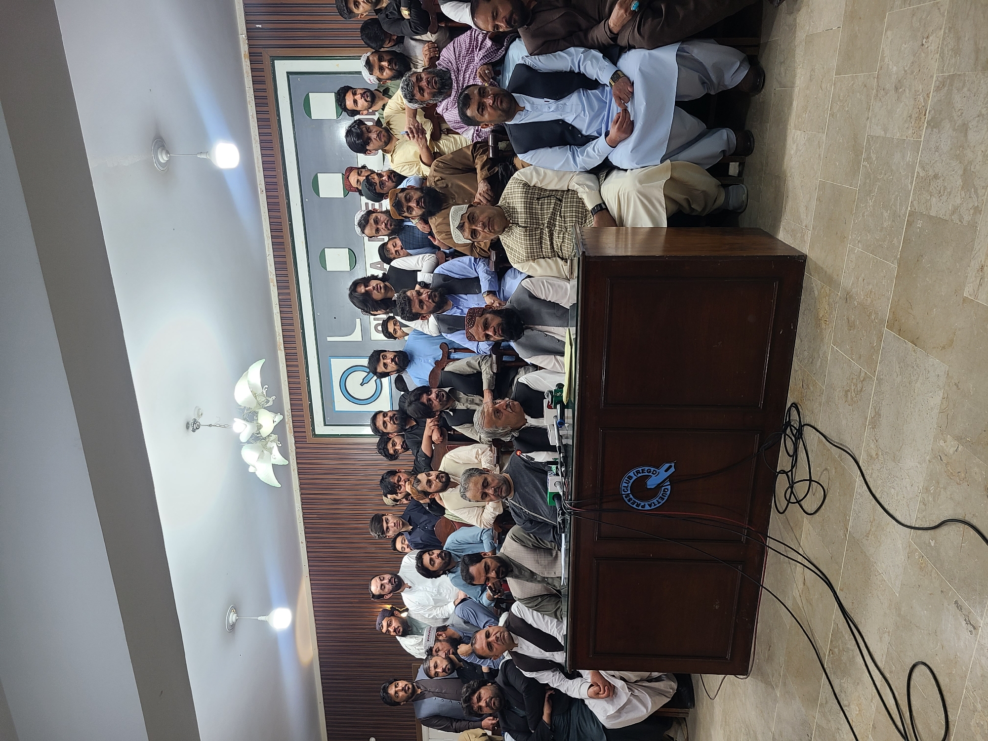 A crowded press conference at the Quetta Press Club in Quetta City Tehsil, Pakistan, features a group of men, primarily wearing traditional attire, seated and standing behind a wooden podium adorned with a blue Quetta Press Club logo. The podium holds microphones and cables. The room has light-colored tile flooring, wood paneling on the walls, and two chandeliers illuminate the scene. A framed logo is mounted on the wall behind the group. The room is full.