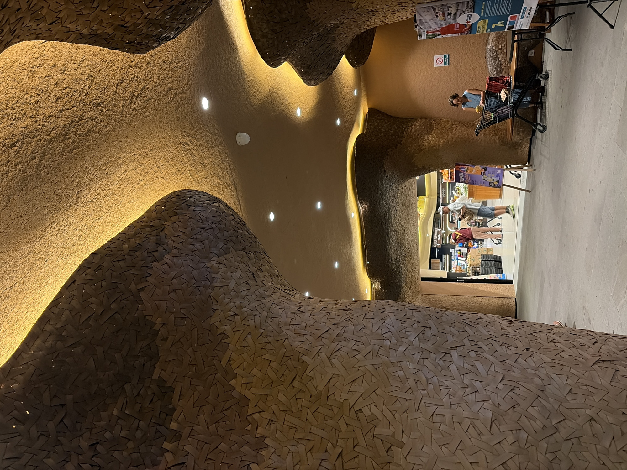 The image depicts an interior scene with organic, flowing architecture and a unique textured wall covering. The primary subject in the foreground is a large, undulating structure made of what appears to be small, interwoven strips of material, possibly paper or wood, in various shades of brown. This textured surface has a light source embedded within it, casting a warm, golden glow on the curved surfaces. The overall architectural style is reminiscent of natural formations, like caves or sand dunes, with soft, rounded shapes and smooth transitions.
In the background, several people are visible, engaged in everyday activities. One woman in a blue top and jeans is standing next to a shopping cart, appearing to select an item. Another person, a man wearing a white t-shirt and shorts, is seen near a display of products. The setting seems to be a commercial space, possibly a market or a store, suggested by the presence of shopping carts, shelves with merchandise, and promotional posters on the walls. One poster has text, but it is illegible. Another small sign reads "NO SMOKING". The lighting in this area is a mix of the warm architectural lighting and the brighter, more functional lighting of a commercial establishment.
The time of day is not explicitly indicated, but the interior lighting suggests it could be evening or a dimly lit indoor environment. The weather is not discernible. The location cues, combined with the stylistic architecture and the general ambiance, hint at a unique commercial or cultural space, possibly a themed area within a larger establishment. The mood is calm and casual, with individuals going about their shopping.