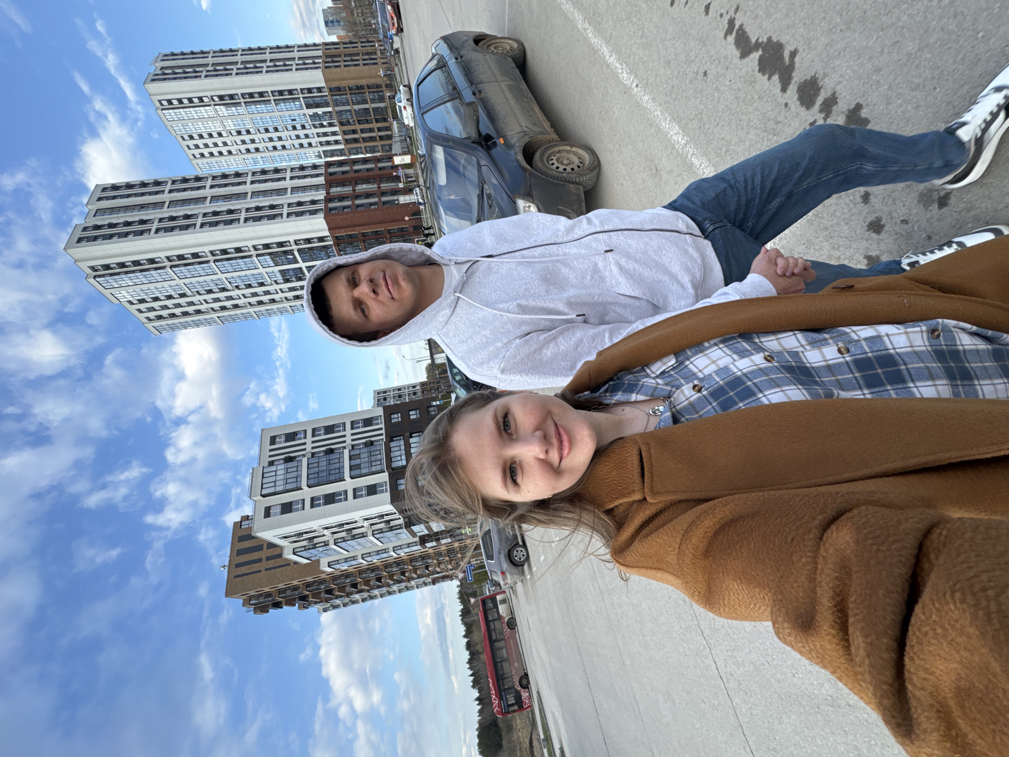 This image, taken in Perm, Russia, presents a tilted, eye-level selfie of a young man and woman. The photograph is rotated 90 degrees clockwise.
The man, positioned to the left (if the image were oriented correctly), has short, dark hair and wears a light grey hooded sweatshirt with a zipper. He looks towards the camera with a neutral yet engaged expression. He is wearing blue jeans and black and white sneakers. His right hand is holding the woman's left hand.
The woman, closer to the viewer on the right, has light brown hair and a warm smile. She is dressed in a blue, white, and brown plaid shirt layered under a textured, warm brown coat. A delicate necklace is visible around her neck.
They are standing outdoors on a wide, paved surface, likely a parking lot or a spacious urban area. The background features several tall, modern multi-story buildings, characterized by numerous windows and a mix of light-colored and brown facades, indicating a contemporary urban or suburban setting. The sky is bright blue with scattered white clouds, suggesting a clear day.
Behind the man, a dark-colored sedan with visibly muddy wheels is parked. Further in the background, a red and white bus can be seen on a road. The overall scene captures a casual, pleasant moment between the two individuals in a bustling urban environment during daylight hours. No discernible text is visible on signs or vehicles.