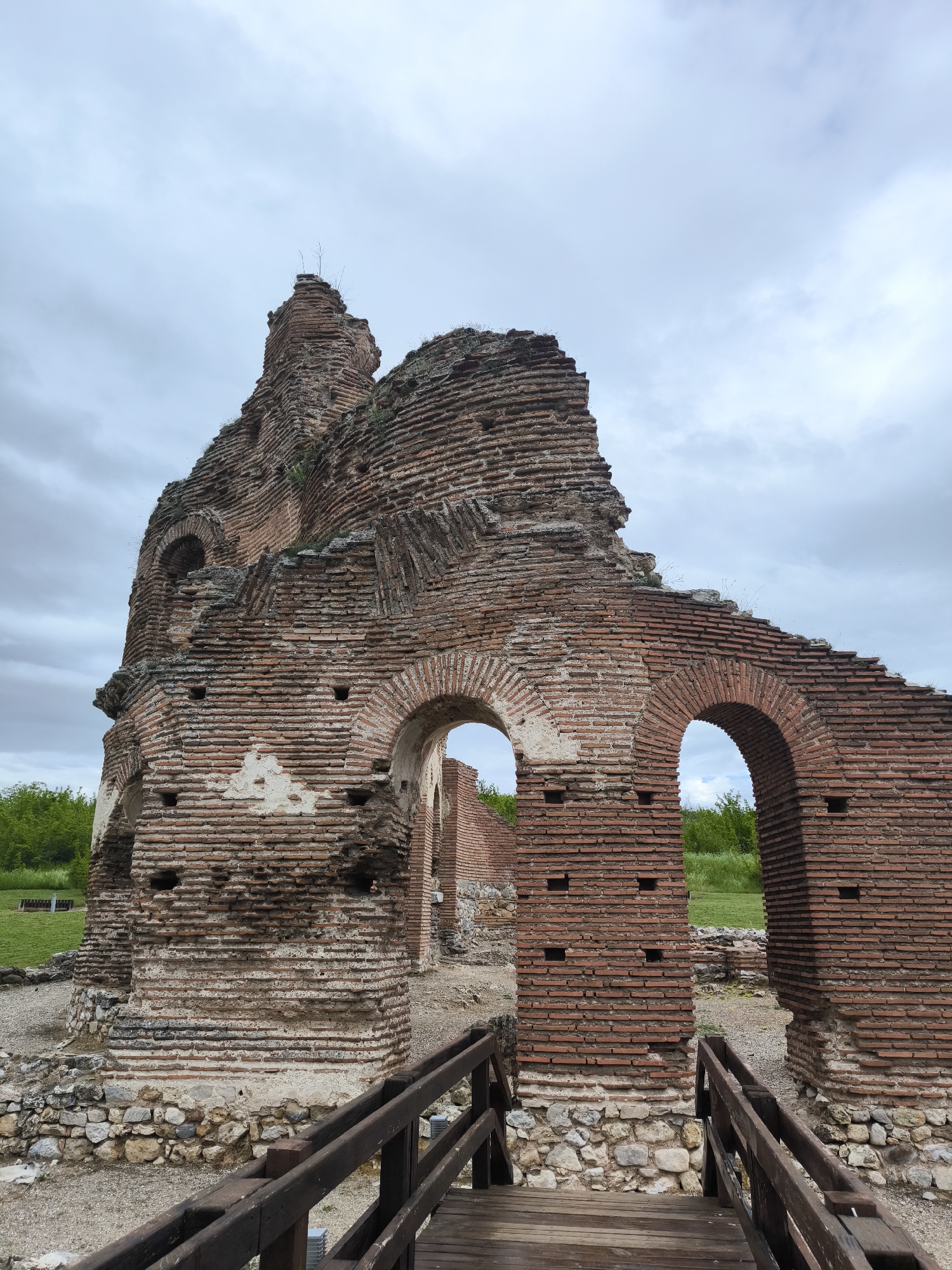 The image shows the ruins of the Red Church in Perushtitsa, Bulgaria. The ruins are primarily constructed of red bricks and stone. A decayed wall with two archways stands in the foreground, with a modern wooden bridge leading towards it. Behind the wall, more ruined structures rise against a cloudy sky. Grass and some trees are visible in the background. No people are visible in the image.