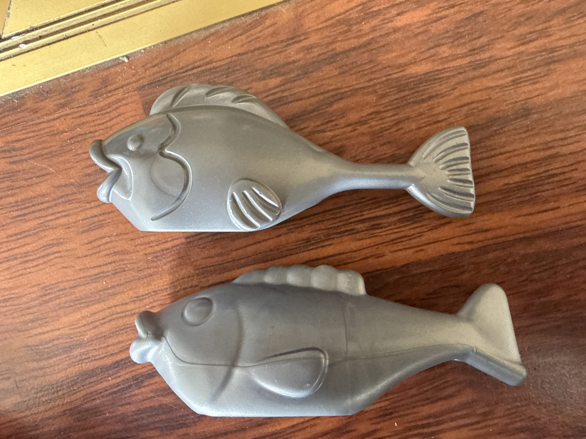 Two gray plastic fish figurines are displayed on a wooden surface. The figurine on the left is positioned vertically, while the figurine on the right is slightly tilted. The wooden surface exhibits a visible grain pattern. The background shows a golden-colored edge, possibly part of a table or frame. There is no indication of location in Ridabu, Norway, or any people present in the image.
