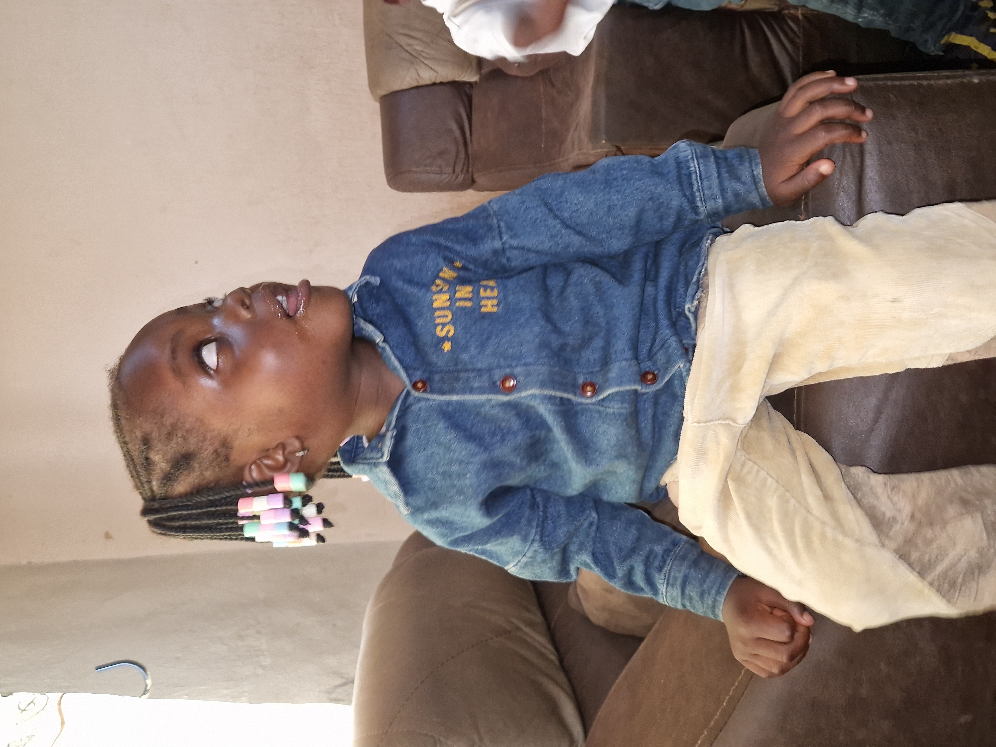 A young Black girl with braided hair adorned with colorful beads sits on a brown leather couch. She is looking upwards and to her right, with her tongue slightly protruding. She is wearing a blue denim shirt with "SUNSHINE" embroidered on it in yellow, and light brown corduroy pants. The setting appears to be an indoor room in Jos, Nigeria, characterized by a plain, light-colored wall and the brown leather couch. A second child, partially visible on the right, is wearing a white shirt. The lighting is natural, coming from the left side of the frame.