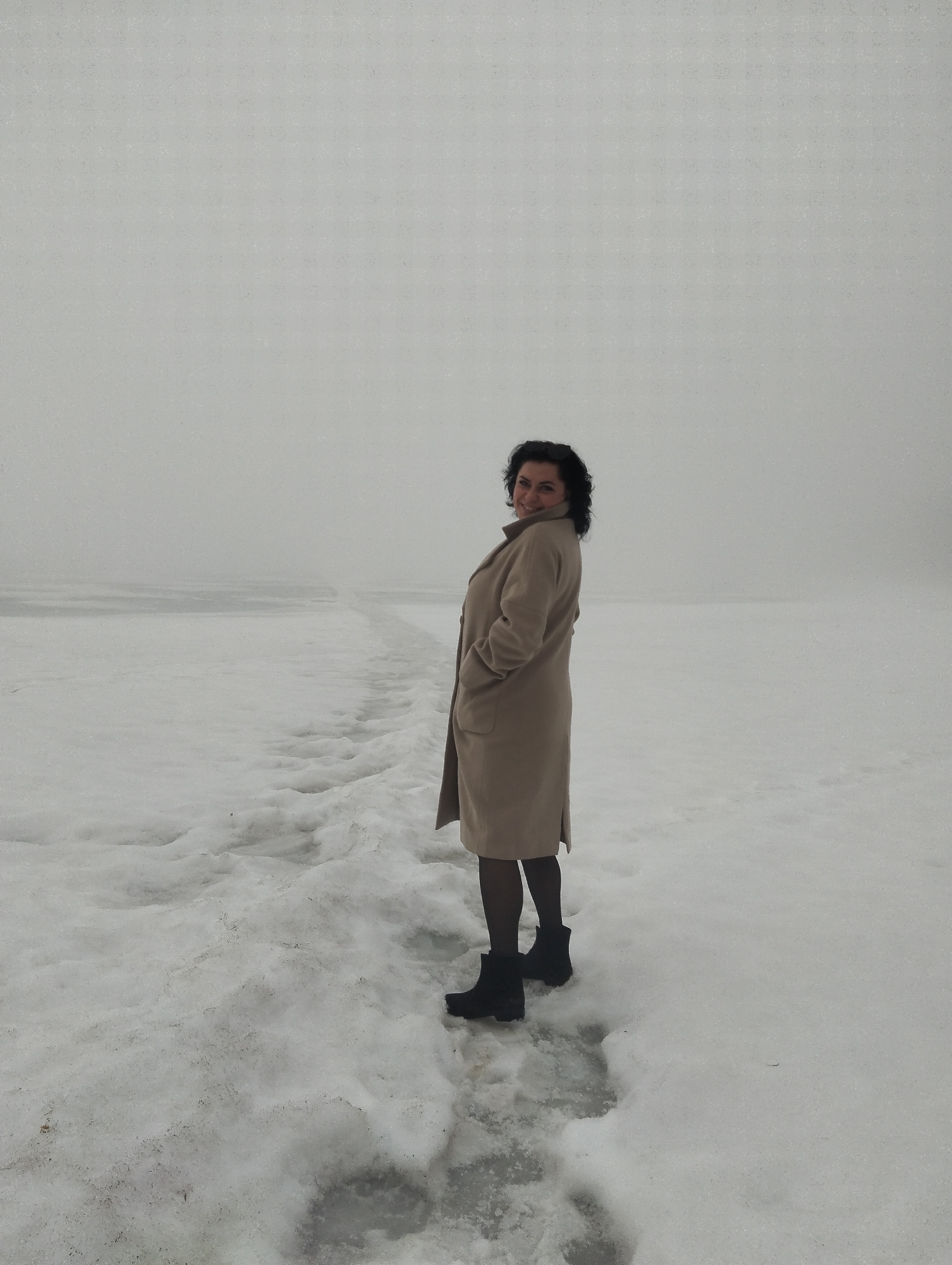 A woman is standing on a snow-covered surface. She is facing away from the camera, but looking back over her shoulder and smiling. She is wearing a long, light-brown coat, black tights, and black boots. The ground is covered in snow, with patches of ice visible. In the background, there is a hazy body of water that fades into the fog. Based on the appearance of the landscape, this could be a snow-covered river or lake in the Votkinsk area, Russia. The pathway of snow in front of her suggests that others have walked on it recently.