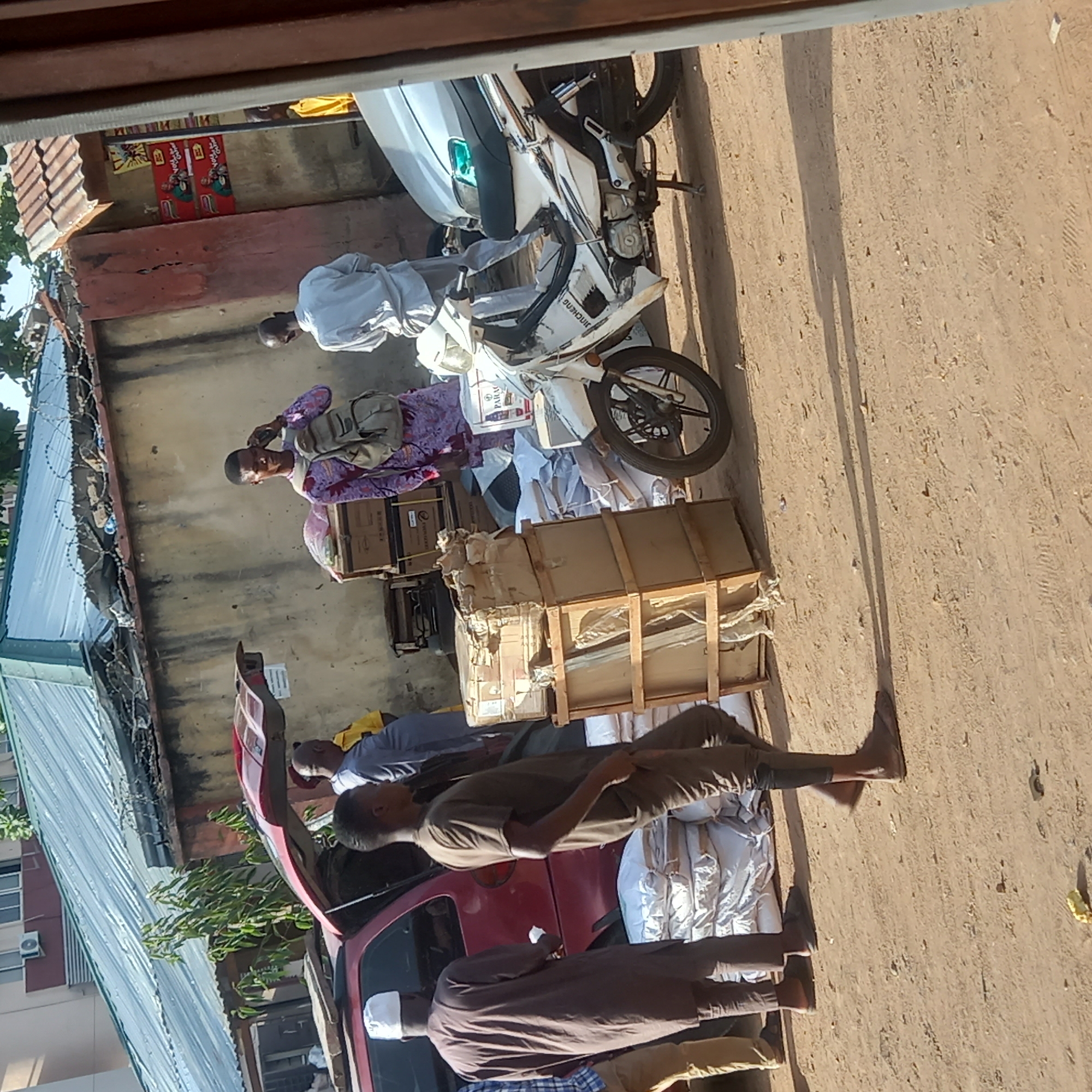 This outdoor scene in Dadi Riba, Nigeria, captures several individuals engaged in what appears to be the handling and transportation of goods on a bright, sunny day. The ground is unpaved, dusty, and shows tire tracks.
In the foreground, a man wearing a brown robe and a white cap stands next to a red car with its trunk open, while another man in a brown t-shirt and shorts stands nearby, looking towards the vehicle. White bundled items are placed on the ground next to the car.
Towards the center, two white motorcycles are parked. One motorcycle clearly displays "JINCHENG" on its side. Around the motorcycles are numerous cardboard boxes and wooden crates, many containing items wrapped in plastic or mesh, suggesting merchandise. A man in a white robe and another individual in a patterned purple shirt carrying a shoulder bag are visible among these goods, seemingly involved in their arrangement or movement. A white bag with "PARAC" visible on it is also near the motorcycles and boxes.
The background features concrete buildings with corrugated metal roofs, some topped with barbed wire fencing. Bright sunlight casts distinct shadows, indicating a clear day. On one building's wall, red posters are visible, prominently displaying "Viju Milk Drink" alongside a cartoon character. The overall scene suggests a vibrant area focused on commerce and logistics.