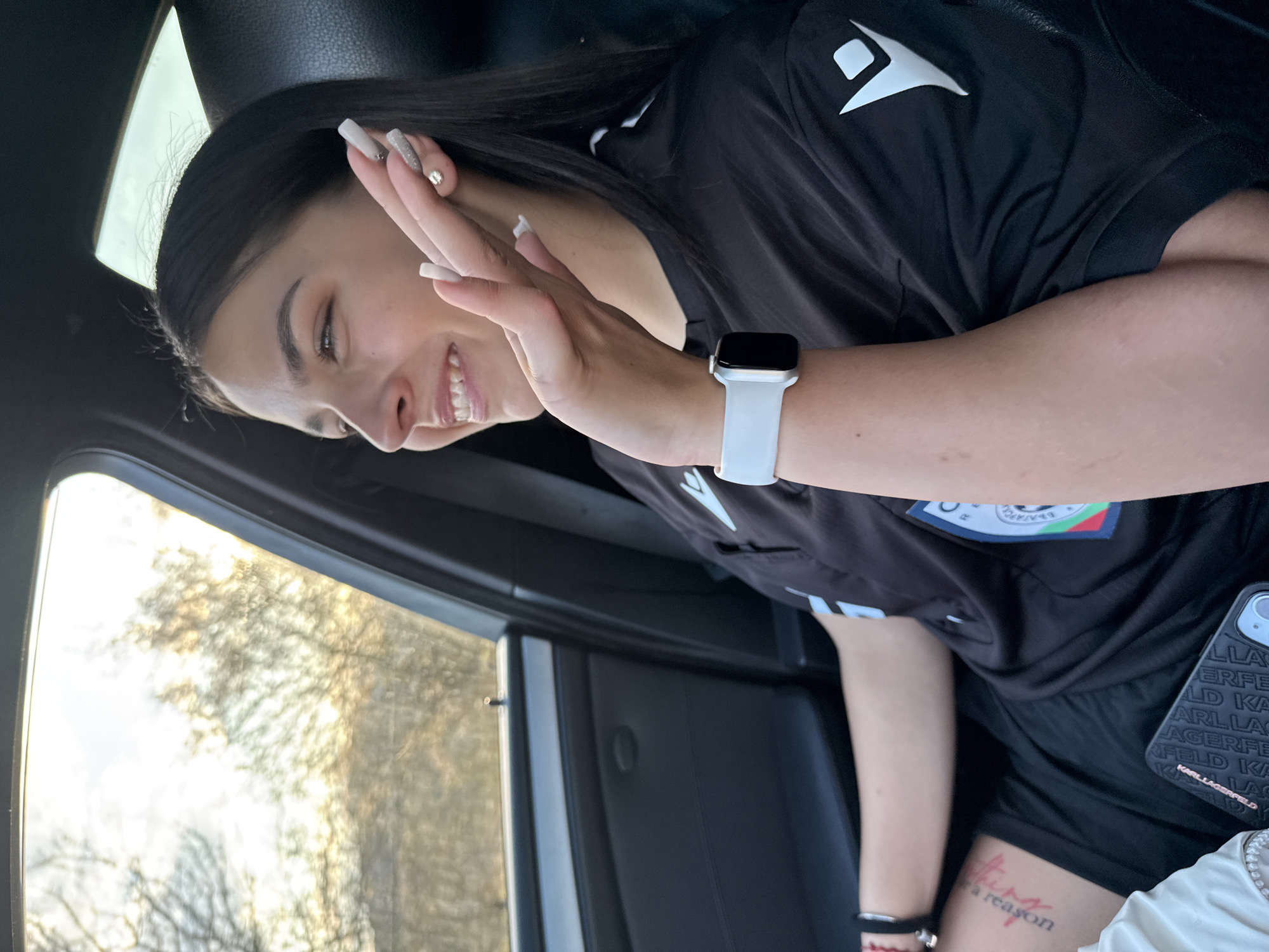 A young woman is seated inside a vehicle, possibly a car, as indicated by the window and interior trim visible. She has dark hair, light skin, and is smiling while raising her hand in a waving gesture. She is wearing a black sports top with a white logo and a white Apple Watch. A phone case with "KARL LAGERFELD" printed on it is visible. Part of a tattoo is visible on her leg. The background visible through the car window indicates a natural environment with trees and sunlight. The location is Gorna Rositsa, Bulgaria, as requested.