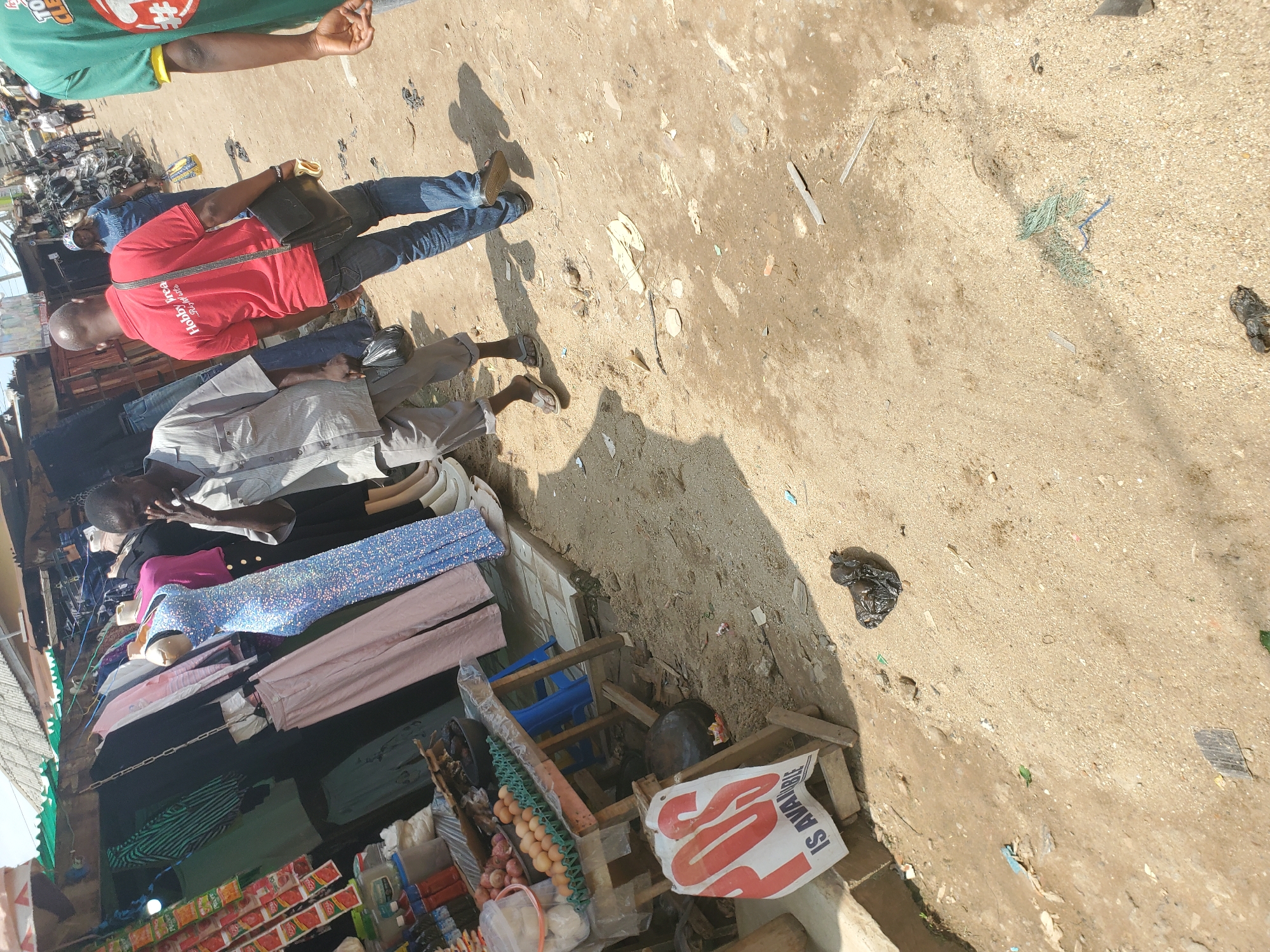 The image depicts a bustling market scene in Shangisha, Nigeria. The primary subjects are people and merchandise. A man in a red t-shirt with the text "Hobbit-Area" and a black bag slung over his shoulder is visible in the foreground. Another person, partially obscured, is wearing a grey and white striped shirt. The ground is a dirt and sandy surface, typical of an outdoor market.
Various goods are displayed, including clothing such as a sparkly blue dress and pink trousers, and a rack of what appear to be condiment packets, possibly for food. There are also eggs in cartons and some round vegetables, likely onions. A wooden cart is laden with these items. A white plastic bag with red text that reads "IS AVAILABLE" is prominently displayed on the cart. The scene appears to be set during the day, with strong shadows indicating bright sunlight. The overall impression is one of everyday commerce and activity in a busy Nigerian market. There is some scattered debris on the ground, including plastic bags and what appears to be netting. The overall atmosphere seems active and possibly crowded, though only a few individuals are clearly visible.