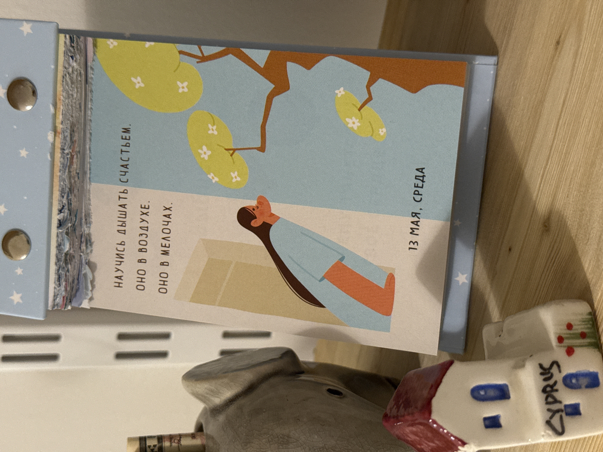 The image shows a desk calendar with a cartoon illustration of a person lying beneath a tree with light green leaves, a blue background, and a brown branch, dated "13 мая, среда" (May 13th, Wednesday) with text in Russian that translates to "Learn to breathe happiness. It's in the air. It's in the little things." The calendar is open and resting against a gray pig figurine and a miniature white ceramic building with a red roof and blue windows, labeled "CYPRUS" in black marker. The setting appears to be indoors, likely a home or office. No location information about Izhevsk, Russia, is present in the image.