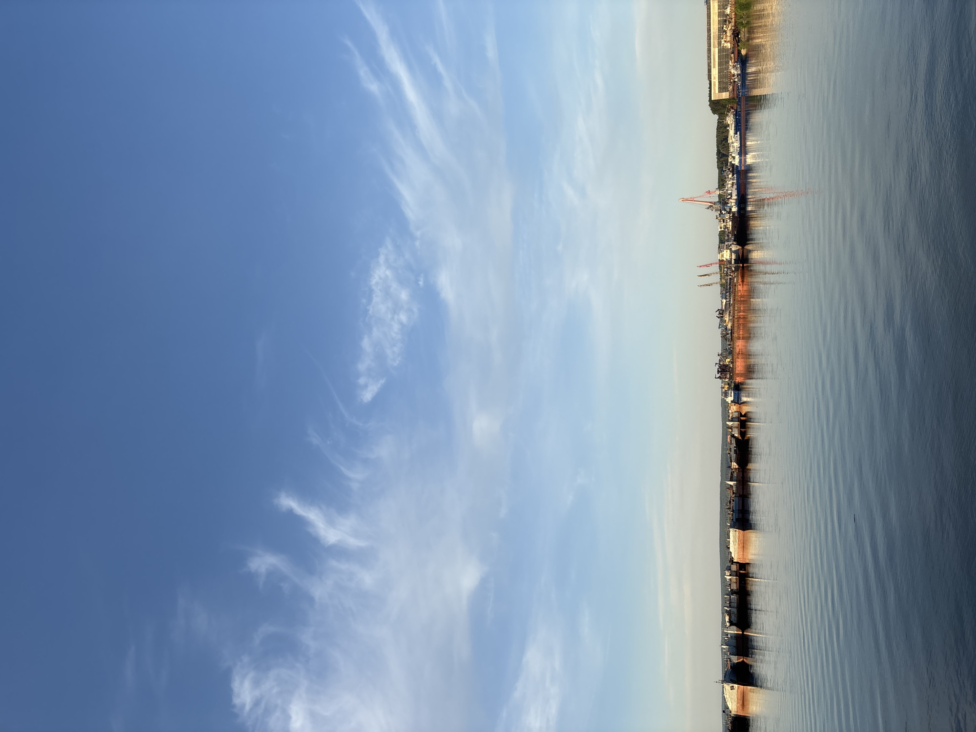 The image shows the Kama River in Chaikovsky, Russia. The right side of the image contains a strip of land with industrial infrastructure, including cranes and buildings, reflected in the river's surface. The sky on the left is blue with wispy clouds.