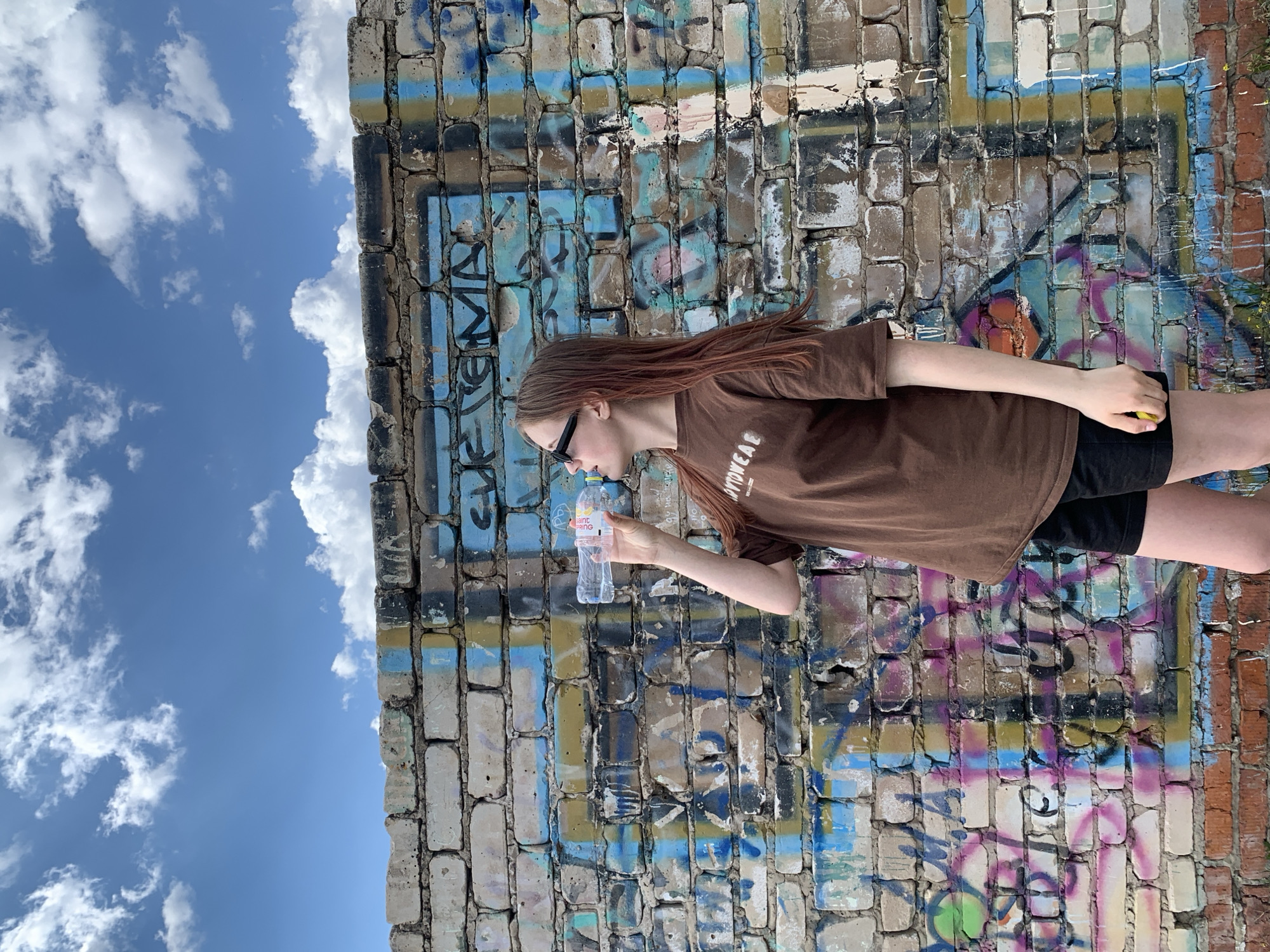 A young person, perceived as a female, is drinking from a plastic water bottle labeled "Saint Spring," with "SPRING" partially visible. She is wearing dark sunglasses and a brown t-shirt with the word "SPORTWEAR" printed on it. She also wears black shorts. The background consists of a textured brick wall covered in graffiti of various colors and patterns. To the left of the wall, there is a blue sky with scattered white clouds. The photo appears to be taken in an urban environment. The location of the scene is Фоки, Russia.