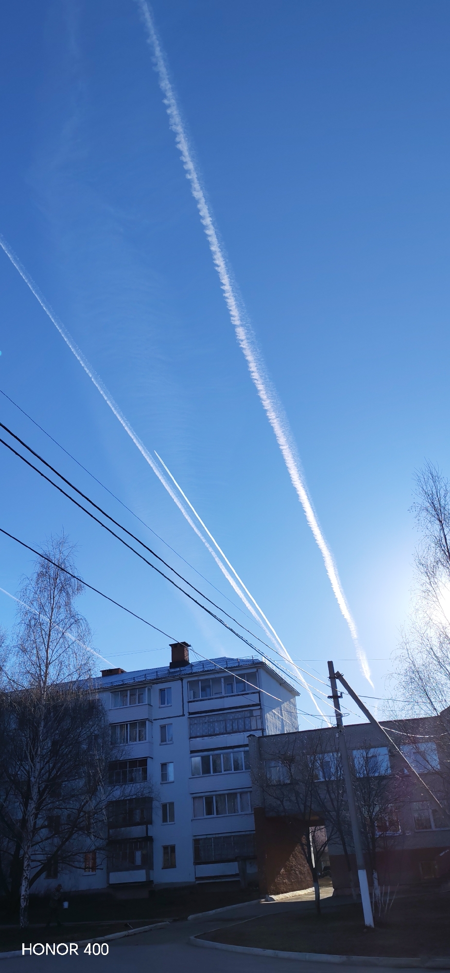 The image shows a bright blue sky with multiple contrails of airplanes crisscrossing above buildings and utility lines in Noviy, Russia. The buildings appear to be multi-story residential structures. Bare tree branches are visible in the foreground, indicating it might be late autumn or early spring. A utility pole stands in front of one building. The bottom left corner shows the branding of the phone used to take the image, a 'Honor 400'. The sun is in the background on the right side of the photo.