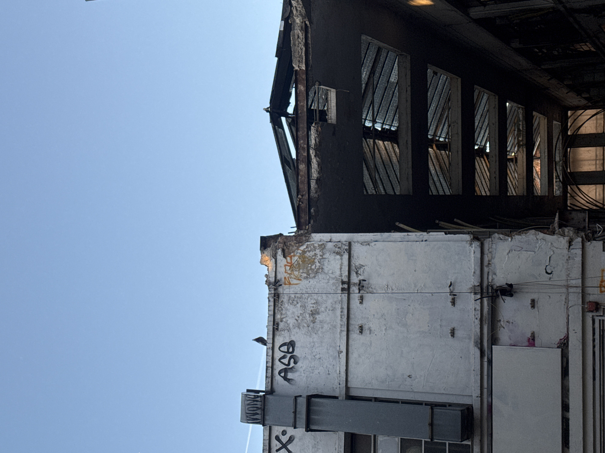 An architectural view from a low angle shows a deteriorated, derelict building in London, United Kingdom. The lower structure features a weathered, white-painted wall with graffiti, including the letters "ASB". A gray duct runs vertically. The upper structure is black, with rows of windows featuring angled metal bars. The sky is clear blue. The overall condition suggests urban decay.