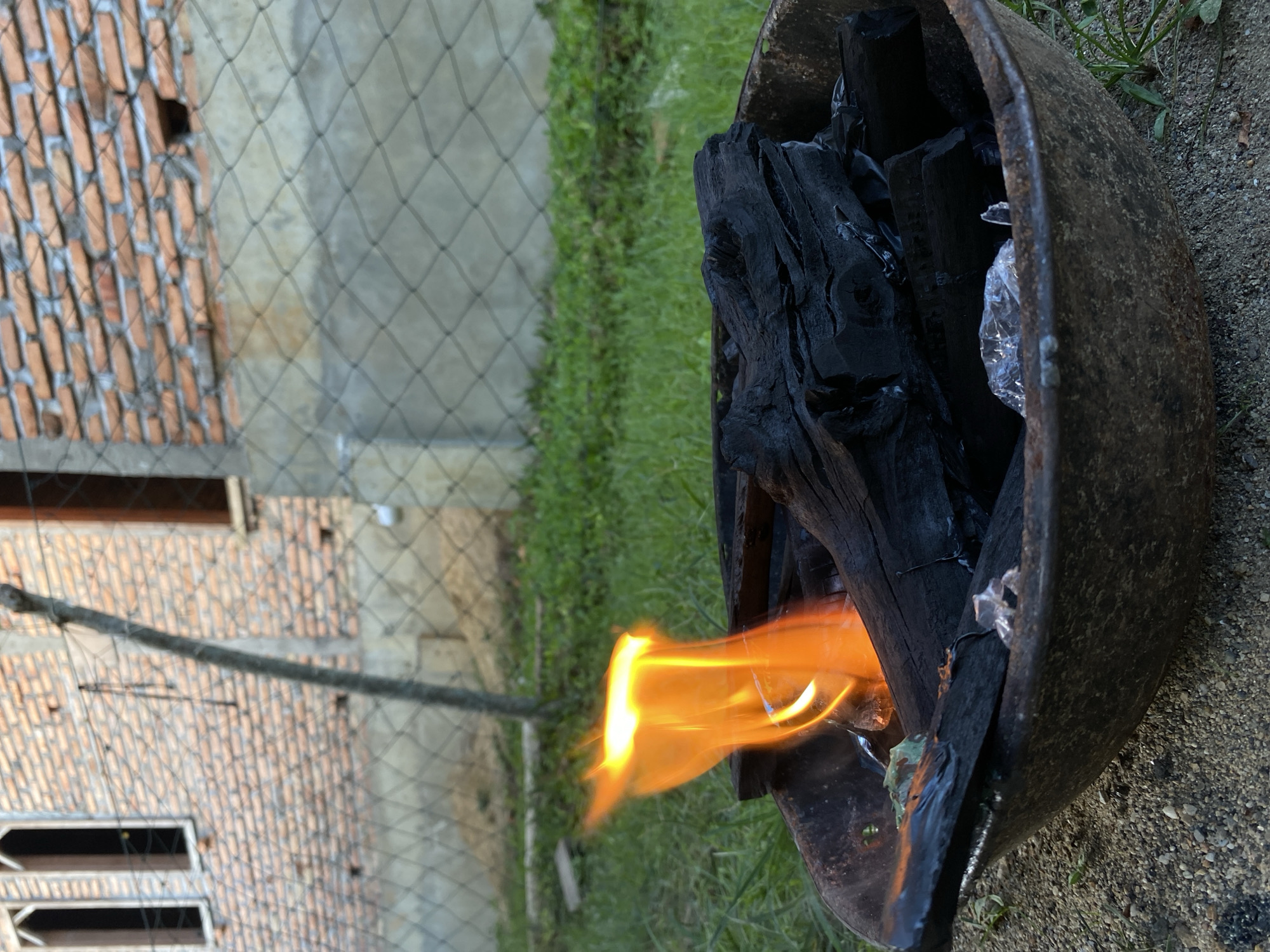 The image depicts a fire burning inside a rusted metal container, likely a makeshift grill or incinerator, situated outdoors in Keudeublang, Indonesia. Blackened charcoal pieces are visible within the container, along with the flickering orange flames. Adjacent to the container is a patch of green grass and, further in the background, a brick building partially obscured by a mesh fence. A thin branch extends across the fence. The ground around the container is sandy or gravelly. No people or animals are present in the frame.