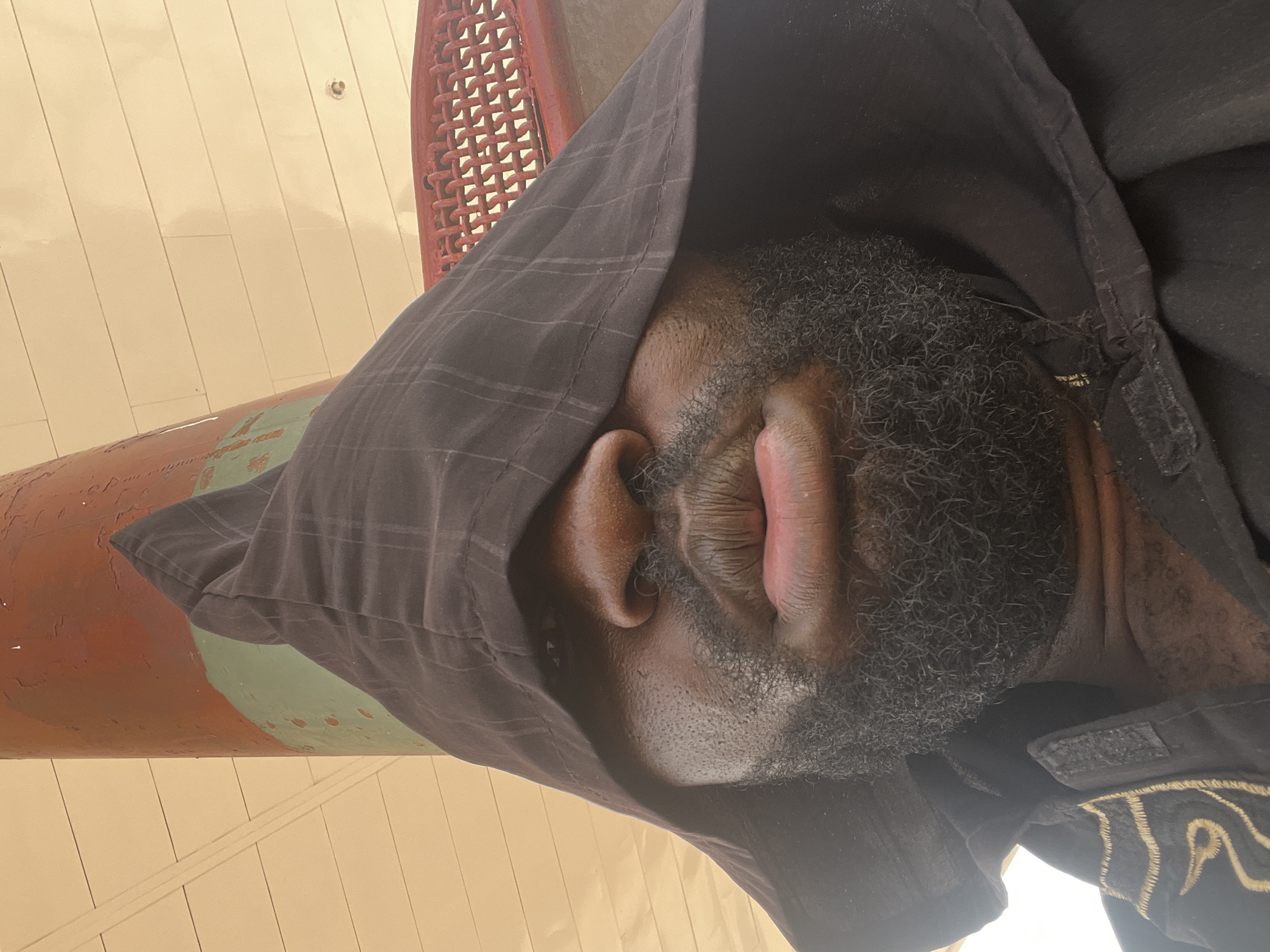 A man is lying down, covered with a dark hooded garment. The location is indicated as Barakin Lerawi, Nigeria. The man has a beard and visible ear. Part of a red object and a light-colored wall are visible in the background. He appears to be resting or asleep.