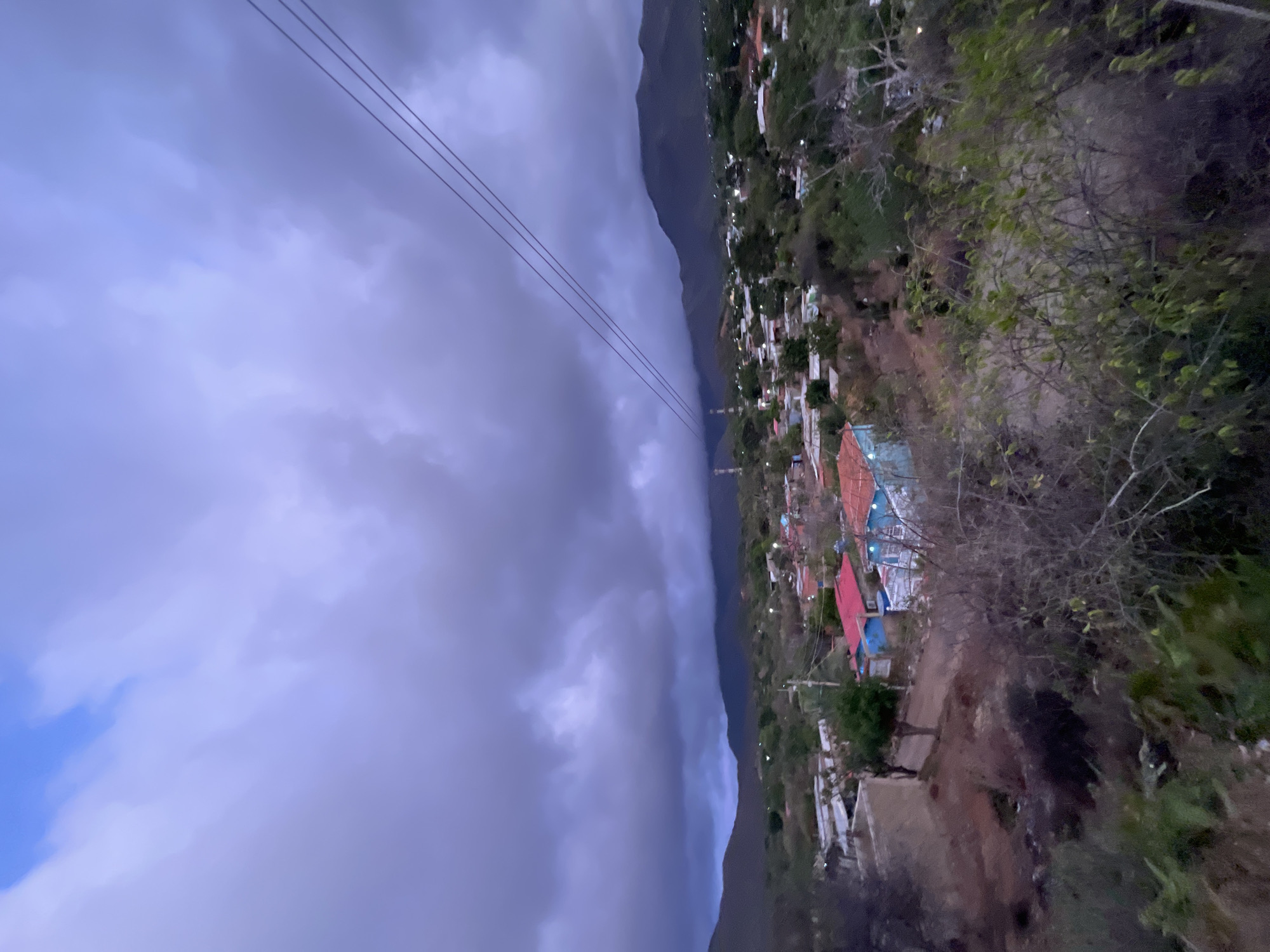 A panoramic view depicts a rural village nestled in a valley, beneath a dramatic, cloud-laden sky. The sky, a mix of deep grays and subtle blues, suggests an approaching storm or the twilight hours. Three taut electrical wires traverse the upper right quadrant of the image, diagonally descending towards the horizon.
The village itself is a cluster of houses with red and blue roofs, interspersed with green vegetation and trees. The terrain is uneven, with dirt paths winding between the structures. Some houses are brightly colored, standing out against the muted tones of the landscape. In the distance, rolling hills or mountains form a backdrop to the scene, shrouded in a light haze. The overall impression is one of a quiet, remote community under a vast and moody sky. The specific location is in Santa Ana, Venezuela.