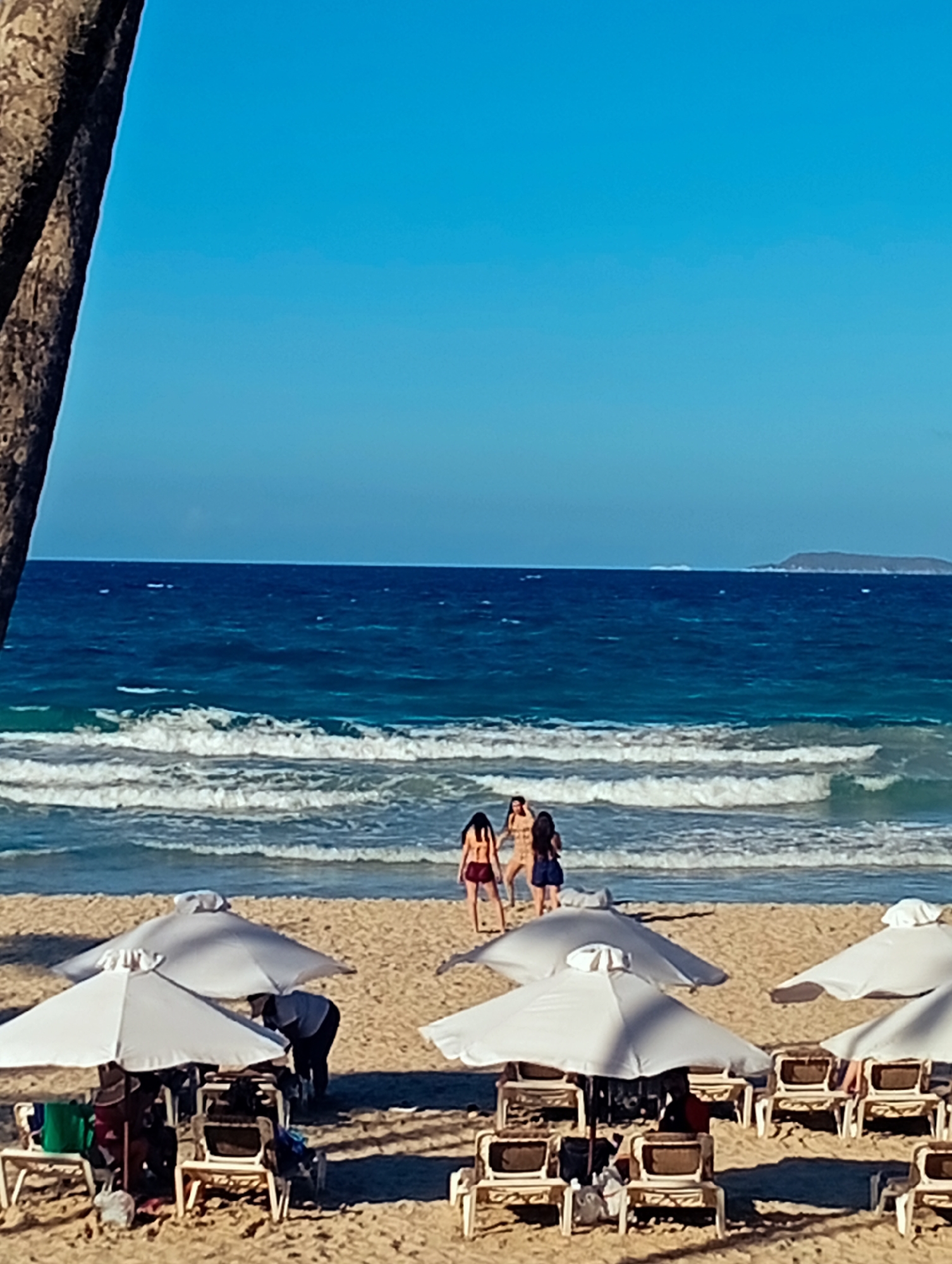The image depicts a beach scene at Playa El Agua, Venezuela. The foreground is dominated by white beach umbrellas and sun loungers, some of which are occupied by people. Beyond this area, the beach stretches towards the ocean. Three women stand near the shoreline, facing the water. The ocean has visible waves and extends to the horizon. In the distance, a landmass is visible. The sky is clear and blue. A palm tree is located on the left side of the photo.