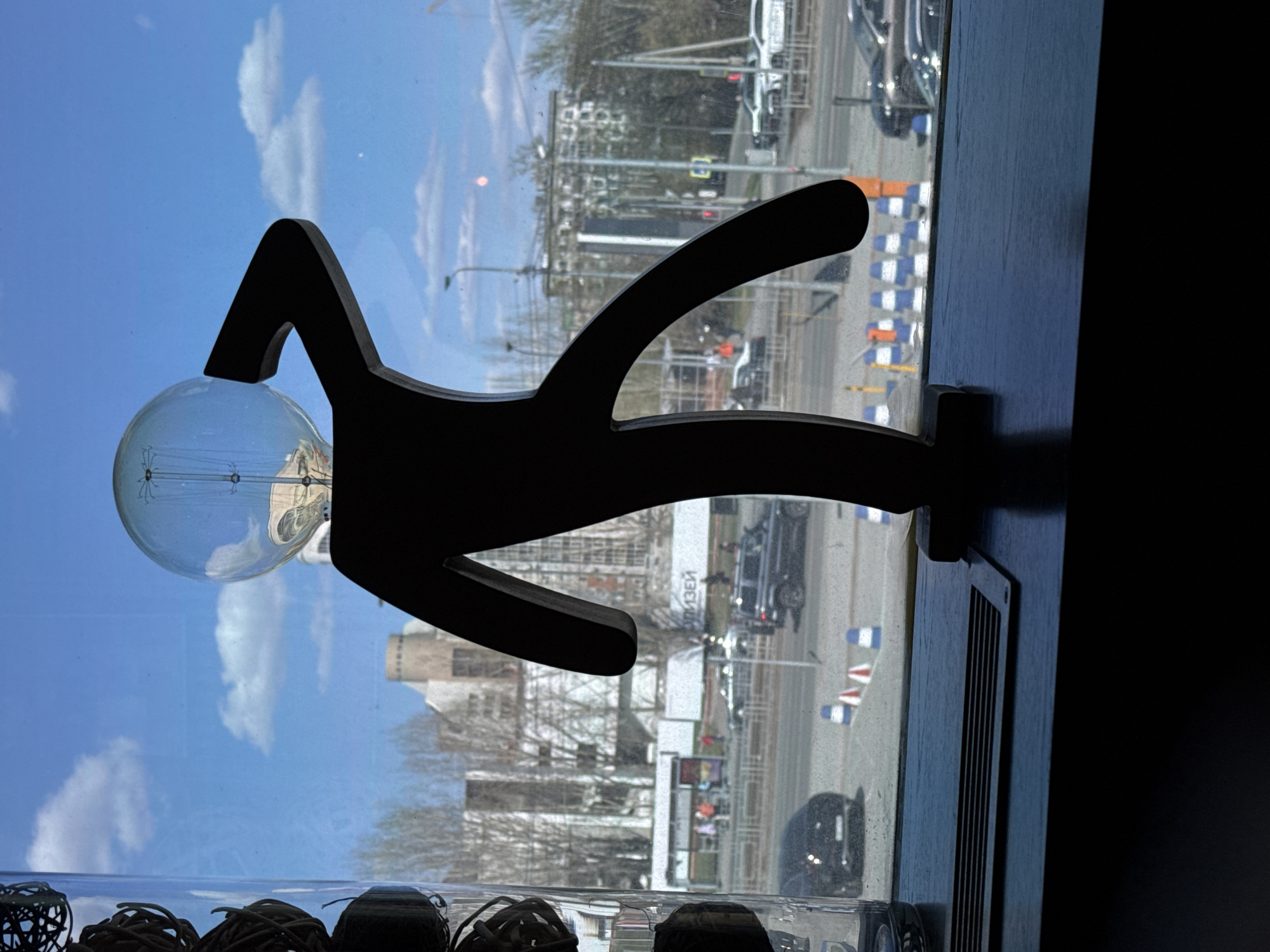 The image shows a stylized, dark silhouette of a human figure with an Edison-style lightbulb for a head, positioned on a dark wooden surface. This object is in the immediate foreground, set against a large window looking out onto an urban landscape.
Through the window, a bright blue sky with scattered white clouds dominates the upper half of the background. The street scene below features various vehicles, including a dark SUV/truck and a dark sedan, some appearing to be moving while others are parked. Traffic lights, streetlights, and power lines are visible, indicating an active urban environment. Bare trees suggest a colder season, such as late autumn, winter, or early spring. In the middle distance, buildings are visible, and a large white sign displays Cyrillic text, which appears to read "МУЗЕИ" (MUSEUM), serving as a significant location cue. Blue and white checkered construction barriers line parts of the street. Reflections of indoor elements, specifically woven wicker balls within a glass container, are discernible on the lower portion of the windowpane. The scene is illuminated by natural daylight, indicating it is daytime. No specific activity or emotion is explicitly conveyed beyond the observational nature of the setting.