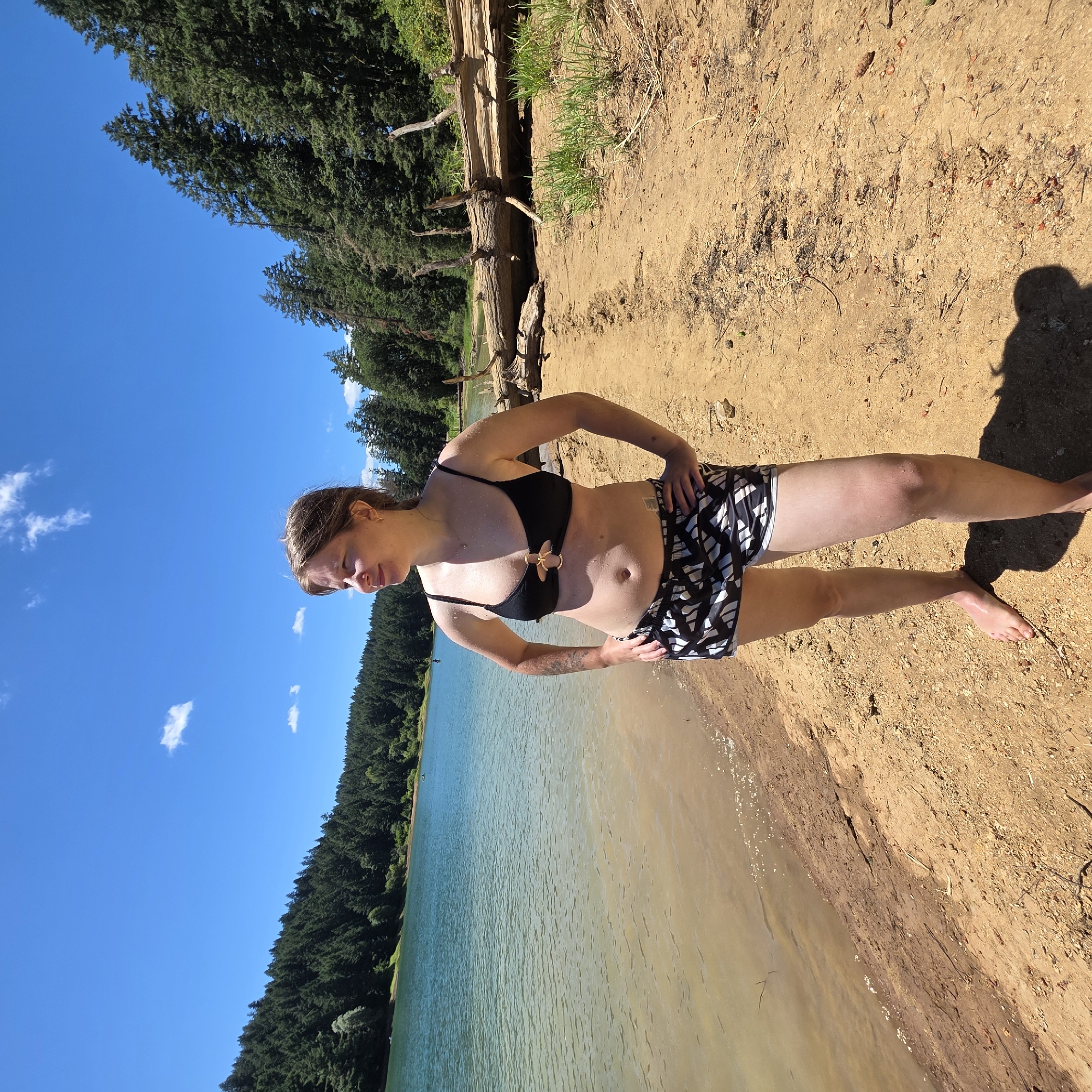 A woman stands on a sandy beach next to a body of water, with a forest of green trees in the background and a clear blue sky overhead. The woman is wearing a black bikini top and black and white patterned shorts. She has her hands on her hips and is looking to her left. The water is a light blue-green color, and the beach is made of light brown sand. There are some fallen trees and branches on the beach behind the woman. The image was taken on a sunny day. The location is a body of water in the city of Cherry Grove, United States.