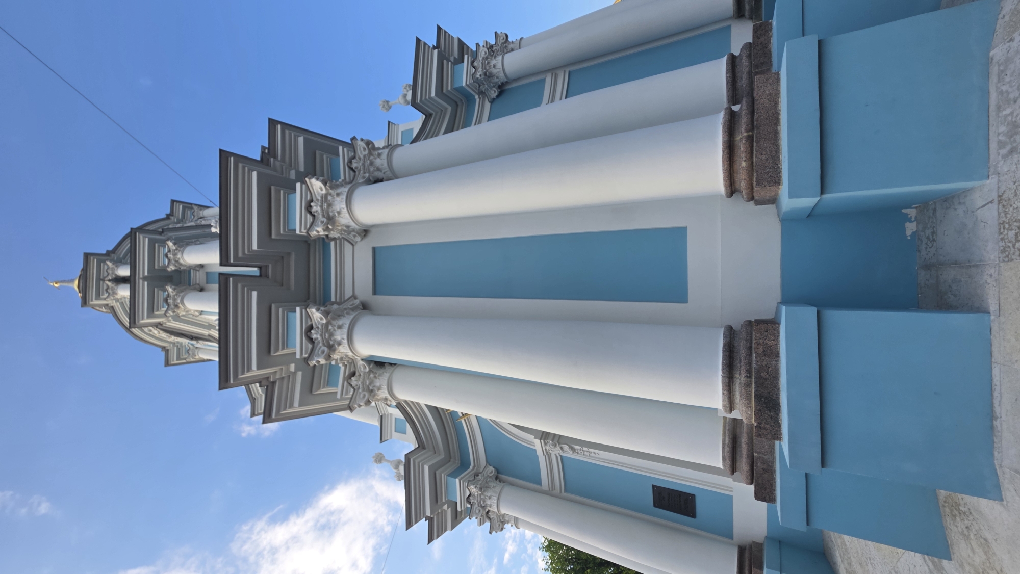This image shows a low-angle view of the Smolny Cathedral in St. Petersburg, Russia. The architecture features a blue and white color scheme, with prominent white columns. The sky is blue with some clouds. The building has ornate details, including classical architectural elements and a golden spire visible at the top. There are several columns stacked upon each other. The base of the structure is finished in granite, and there is a small plaque on one side of the structure. A thin wire is visible against the sky, likely a power line. The image is oriented vertically.