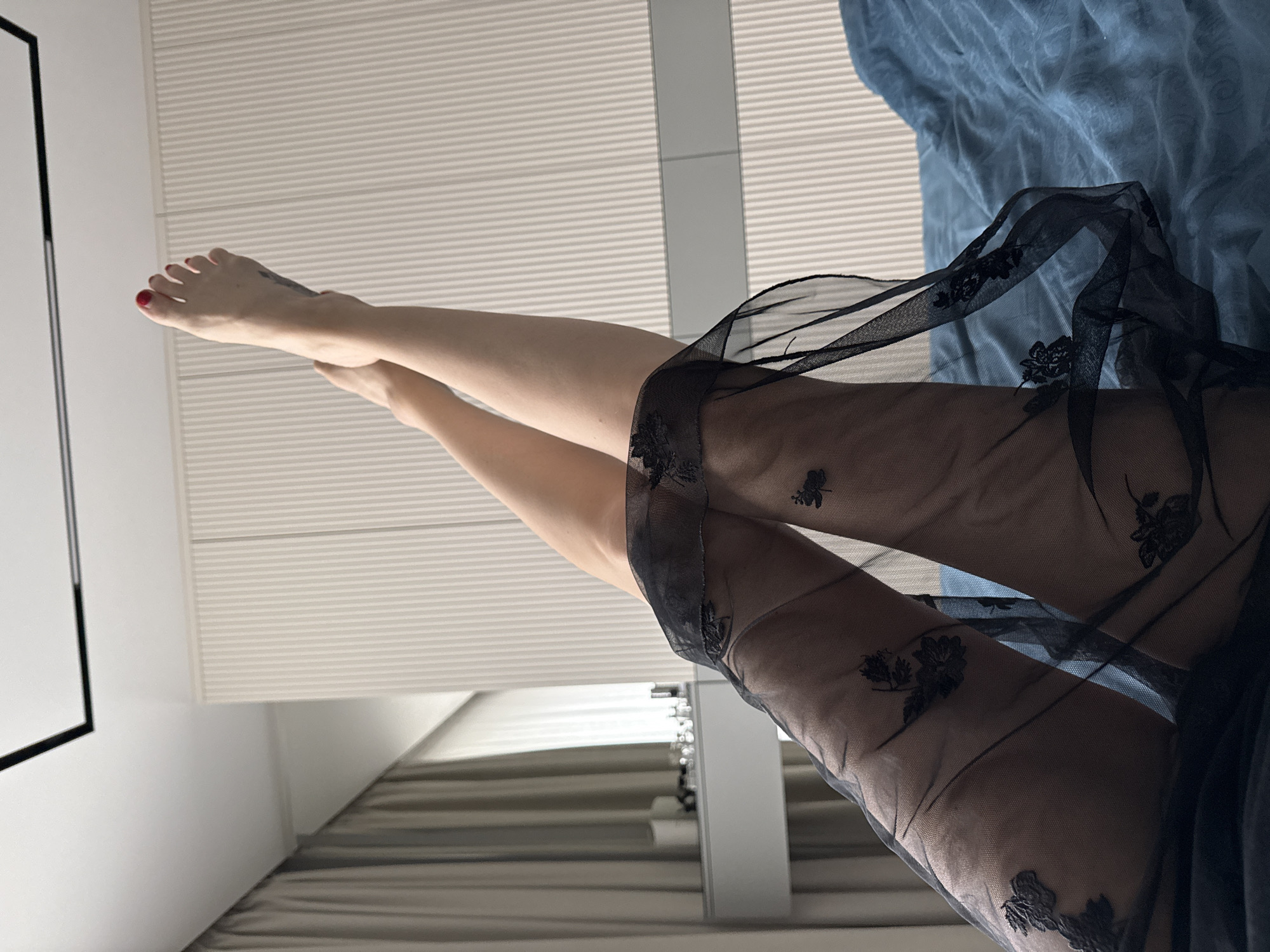 A woman is lying on a bed covered with blue sheets, wearing a black, floral-embroidered, sheer negligee. Her legs are raised in the air. She has red nail polish on her toes. The room has white blinds and a framed object on the wall. The location is Khimki, Russia.