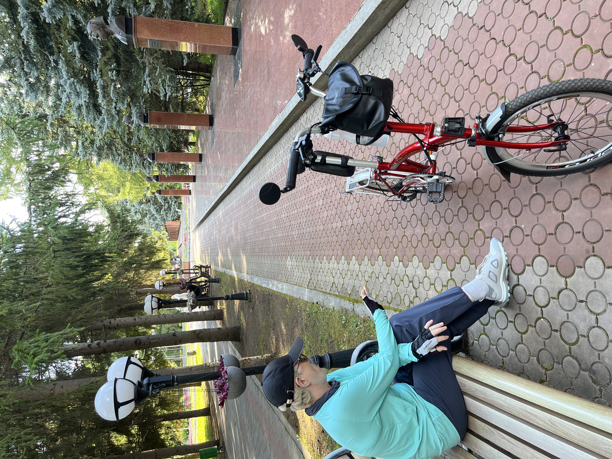 A woman wearing a black baseball cap, sunglasses, a turquoise long-sleeved shirt, dark pants, and gray athletic shoes sits on a wooden bench in an outdoor park. She is holding a smartphone in her left hand and looking down at it. Her right hand is extended, palm up, as if gesturing or holding something. Next to the bench, a red folding bicycle is parked, equipped with a black handlebar bag and a water bottle holder. The park features a paved pathway with an intricate circular pattern, lined with trees and lampposts. In the background, there are busts on pedestals and more trees. The scene is illuminated by natural light, suggesting daytime. This location is identifiable as the Alley of Glory in Almetyevsk, Russia, a park dedicated to honoring notable individuals.