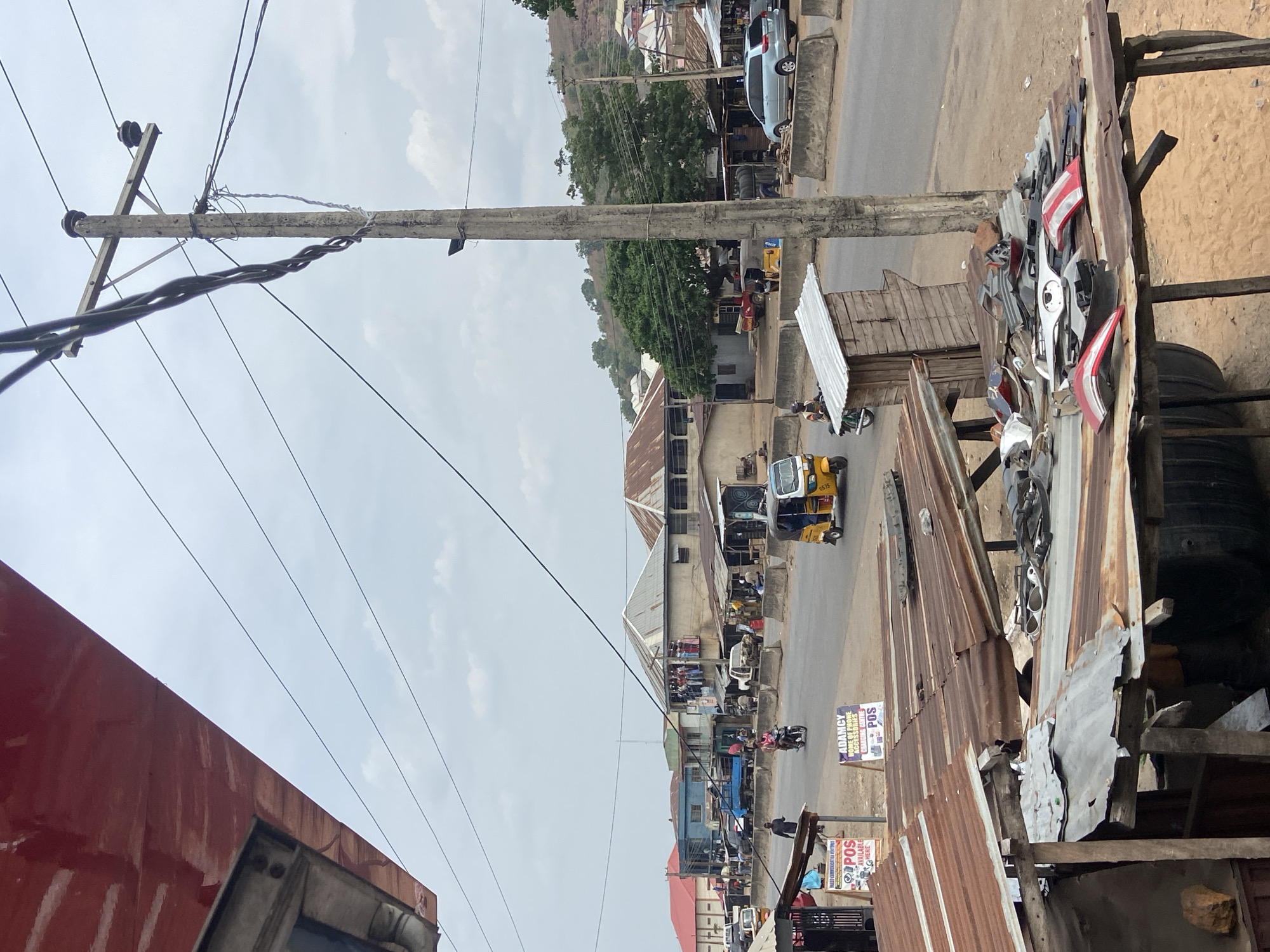 This image captures a bustling street scene, likely in an urban or semi-urban area of Lokoja, Nigeria. The sky is overcast, suggesting a cloudy day or bright haze.
In the foreground, to the left, a large, rustic red surface, possibly part of a building or truck, partially obscures the view. Overhead, several power lines extend across the frame, connected to a prominent wooden utility pole equipped with crossarms and insulators.
The middle ground features a busy street lined with various buildings, many characterized by their corrugated iron roofs, some showing rust and wear. A light-colored car is parked on the right side of the road, while several vehicles, including motorcycles and at least two yellow tricycle auto-rickshaws (Keke Napep), are visible on the street. Numerous people are scattered throughout the scene, walking or engaged in daily activities.
To the right, a makeshift stall or shop constructed from corrugated metal and wood displays an assortment of vehicle parts, including headlights, taillights, and body panels, indicating an auto repair or spare parts business. Further down the street, more such stalls and buildings contribute to the commercial atmosphere. A hill covered with sparse greenery rises in the background.
Visible text on signs includes "ADAMU" or "ADAMC" (likely a business name), "TYRES," "RIMS," "ACCESSORIES," and prominently, "POS AVAILABLE HERE," indicating point-of-sale services. The overall impression is one of everyday commerce and transport in a lively community.