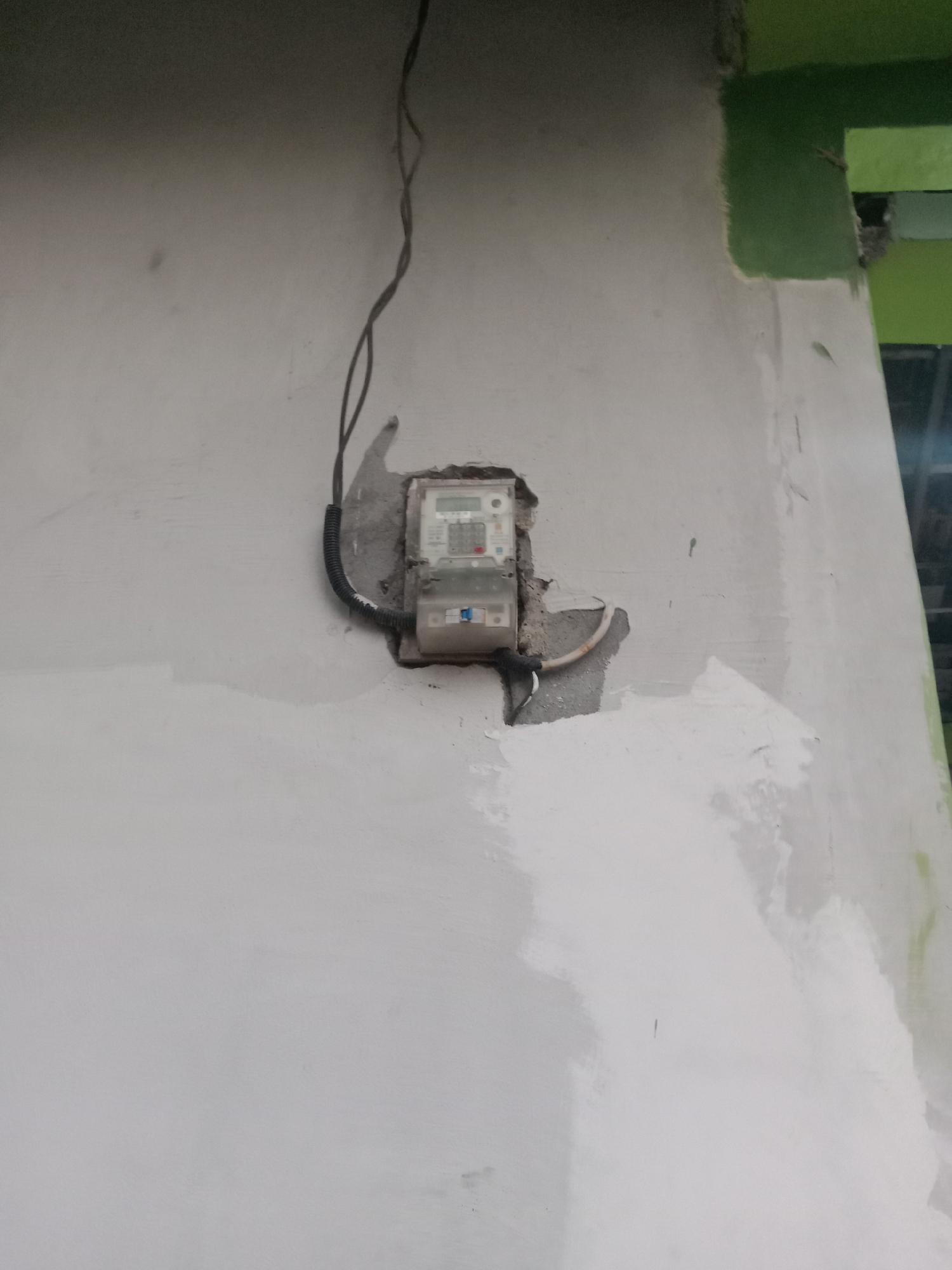 A close-up, eye-level shot shows a white stucco wall with a recessed electrical meter box. The meter box, which is a light gray plastic, has a digital display and some buttons. Two wires, one black and one white, emerge from the top of the meter box, and a black corrugated conduit snakes down from the left side, entering the box. The stucco around the meter box is chipped and rough, suggesting recent work or damage. The top right corner of the image shows a section of wall painted bright green. The overall lighting is dim, with a gradient from dark at the top to lighter towards the bottom, indicating an indoor or shaded outdoor location. There are no people or animals visible in the frame. The location is stated to be Jumput, Indonesia.