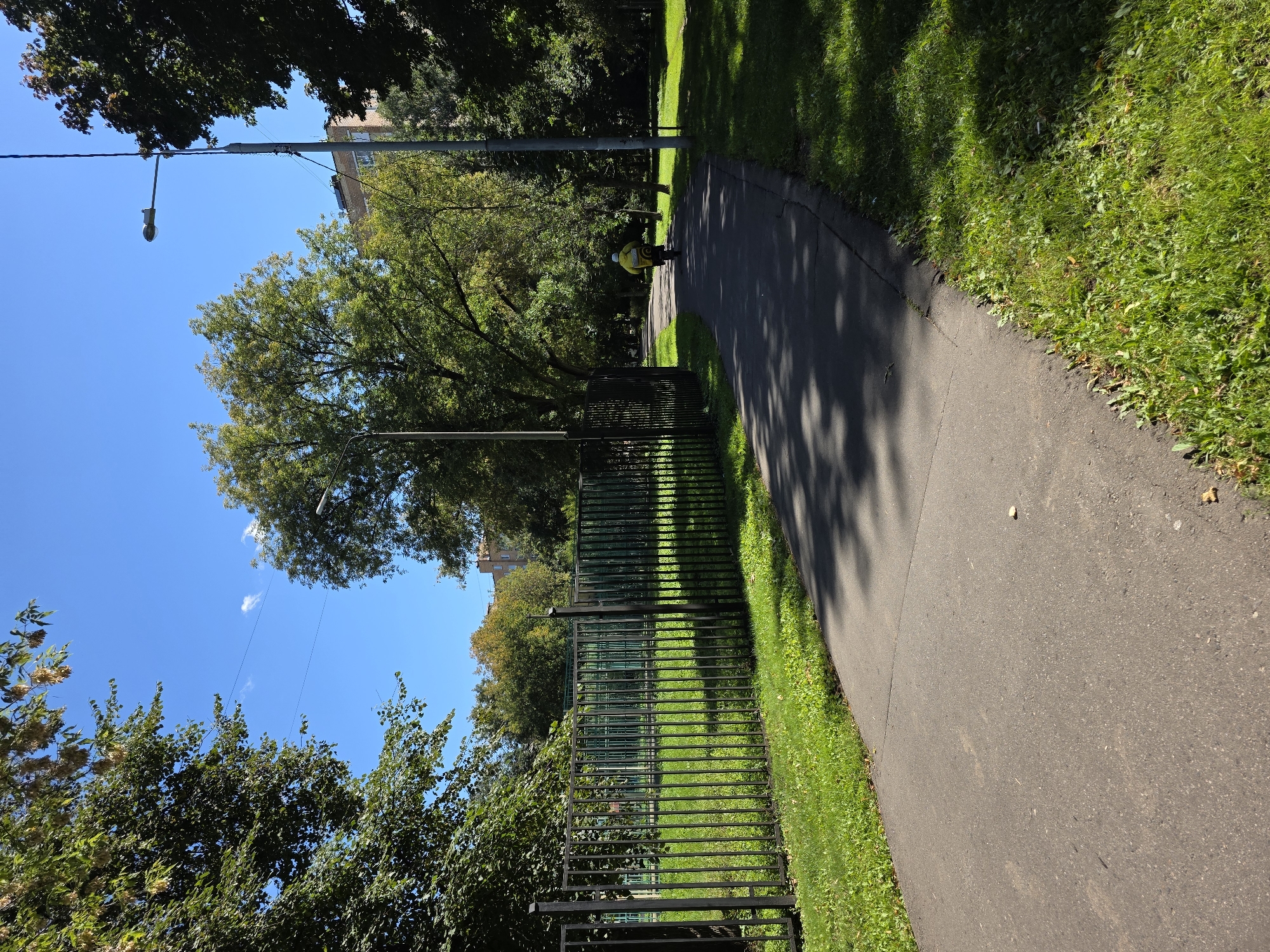 The image shows a paved pathway through a park in Moscow, Russia, on a sunny day. A person in a bright yellow jacket is riding a bicycle away from the camera, down the path. The pathway is bordered by green grass and a black metal fence on the left, with a fenced-off area of grass and trees behind it. On the right, there is more grass and trees, with a tall lamppost and a building visible in the background. The trees are lush and green, suggesting it is summer or early autumn. The sky is a clear blue.
