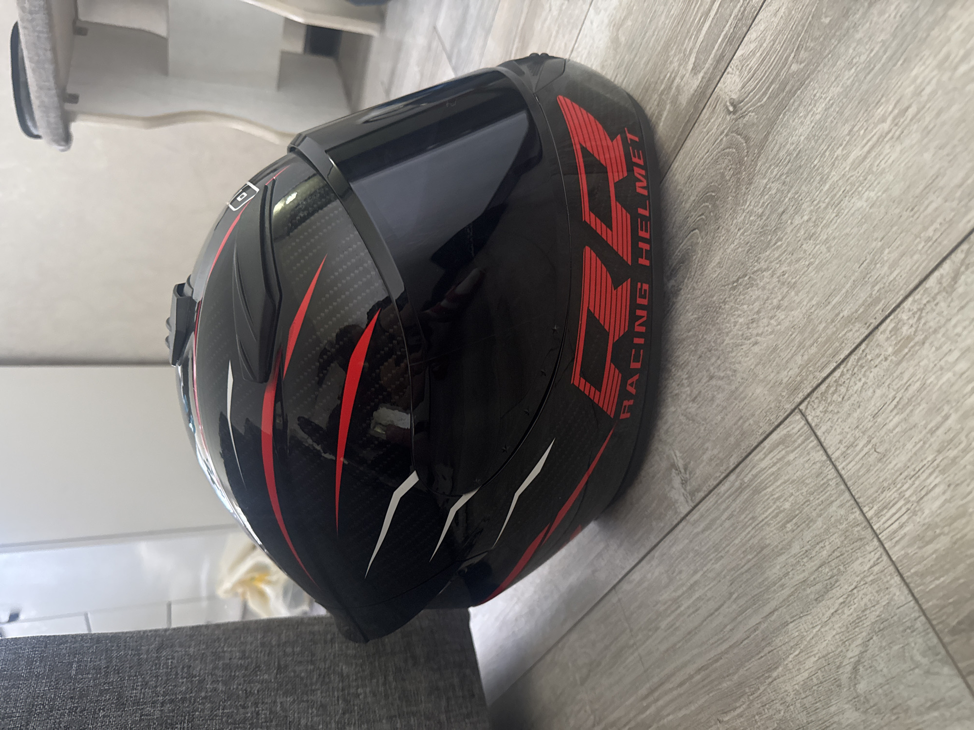 A black racing helmet with red and white accents sits on a light gray wooden floor. The helmet has a carbon fiber-like pattern with red stripes and white accents on the sides. The text "RR RACING HELMET" is visible in red on the bottom of the helmet. A small black object, possibly a camera mount, is attached to the top of the helmet. In the background, a gray textured sofa is partially visible on the left, and a white piece of furniture with carved legs is on the right. The environment appears to be indoors, possibly a residential setting.