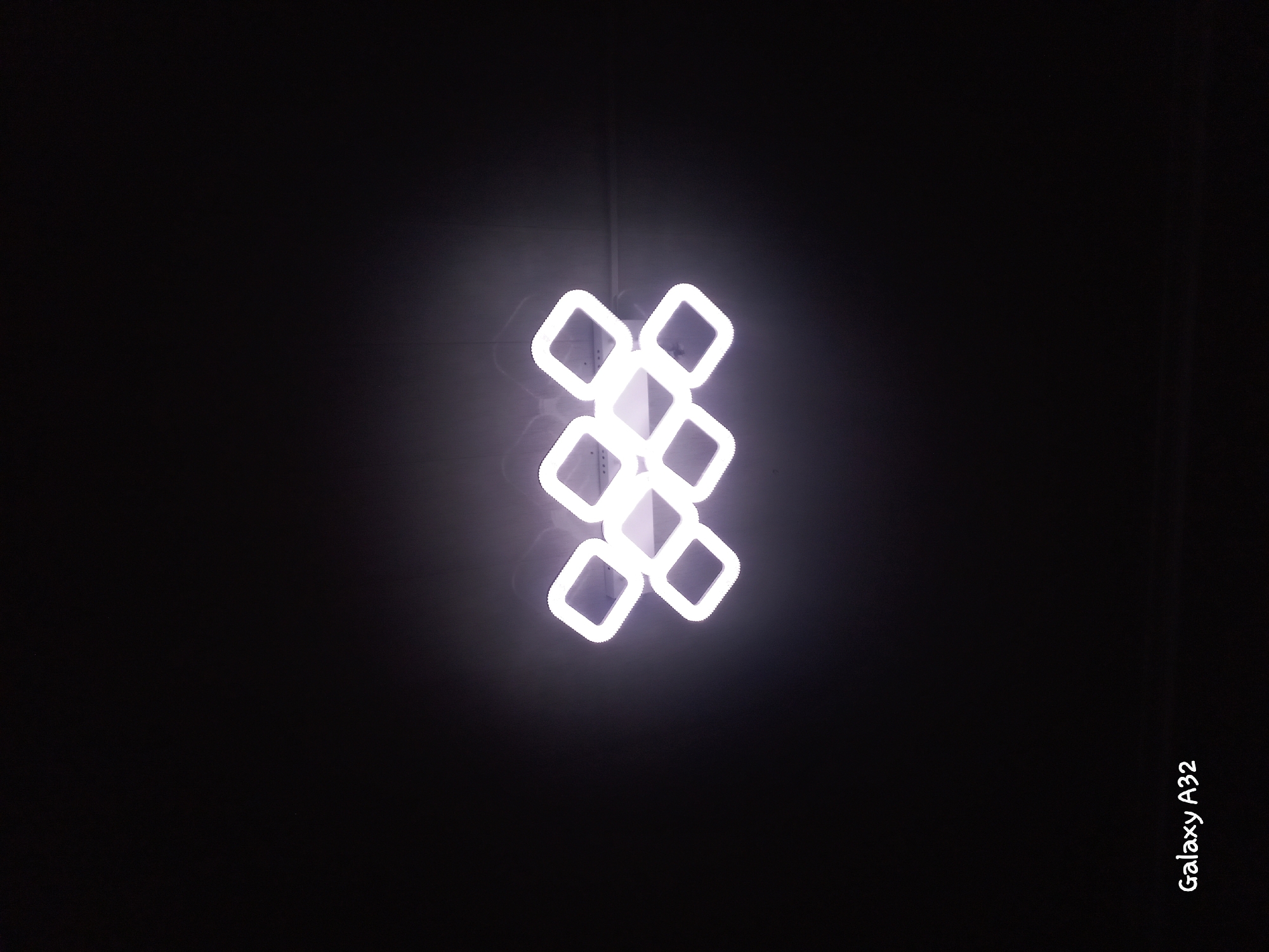 An abstract modern light fixture, composed of multiple square frames arranged in a diamond pattern, illuminates a dark ceiling. The light is a bright, cool white with a slight purple hue, creating soft shadows and a subtle glow. The fixture is centrally located in the frame, against a plain, dark background that suggests an indoor setting, possibly a room or a hall. The text "Galaxy A32" is visible in the bottom left corner, indicating it is a photograph taken with a Samsung Galaxy A32 phone. The location of Kemin, Kyrgyzstan, is mentioned, but the image itself provides no visual cues to confirm this specific location.