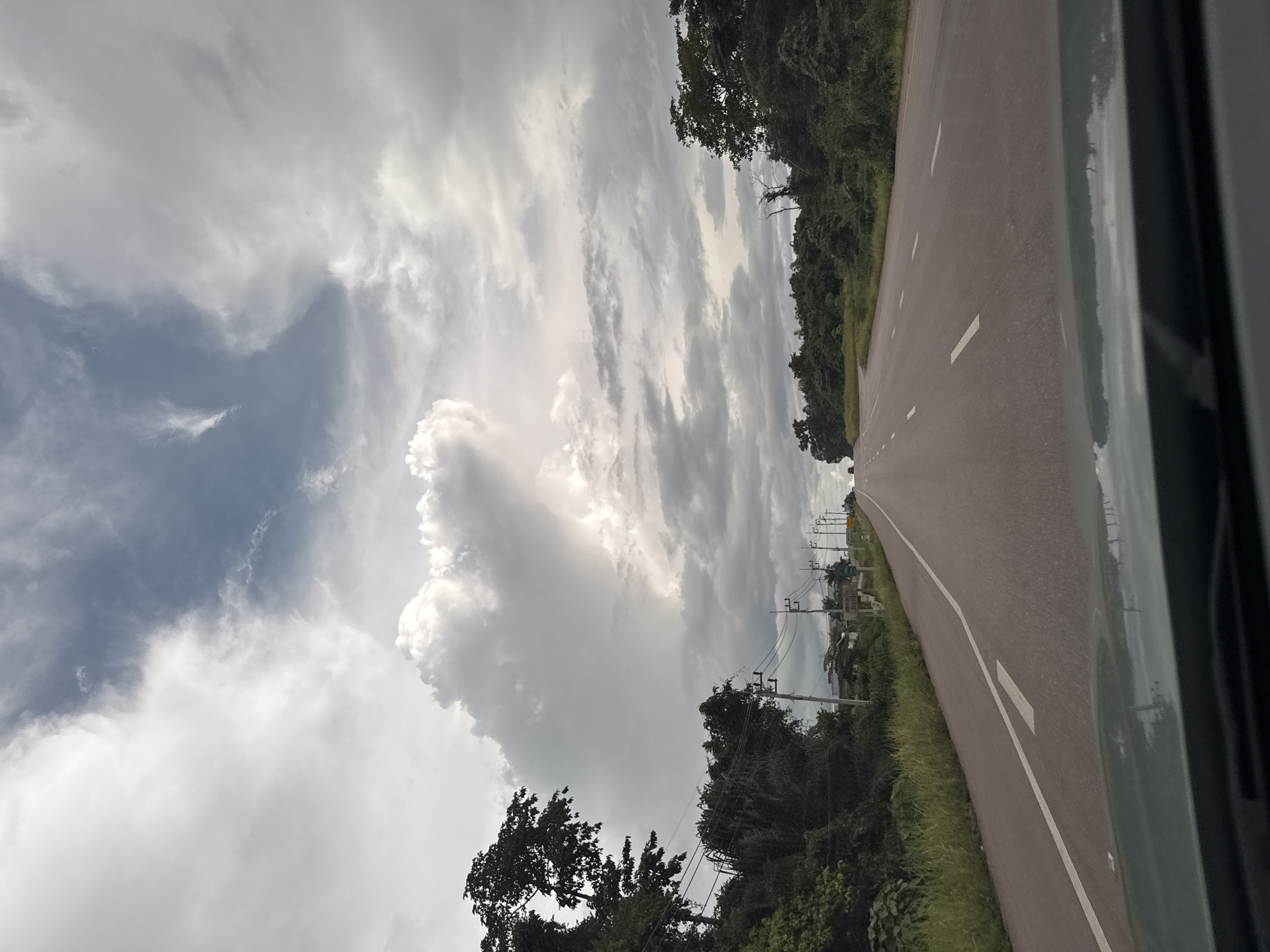 The image captures a view from a vehicle on a two-lane asphalt road, likely in Plai Phraya, Thailand, as suggested by the presence of tropical foliage and a cloudy, overcast sky characteristic of the region. The road stretches ahead, curving gently to the right in the distance. White dashed lines mark the lanes, and a solid white line is visible on the left side. The surrounding environment is lush with dense green vegetation, including tall trees lining both sides of the road. Utility poles with numerous wires are visible along the left side of the road, indicating infrastructure in the area. The sky is filled with dramatic, heavy clouds, with patches of brighter light suggesting breaks in the overcast conditions. A few distant vehicles are visible on the road. The foreground shows the dashboard and windshield of the vehicle from which the photo was taken, with reflections of the sky and road visible on the surface.