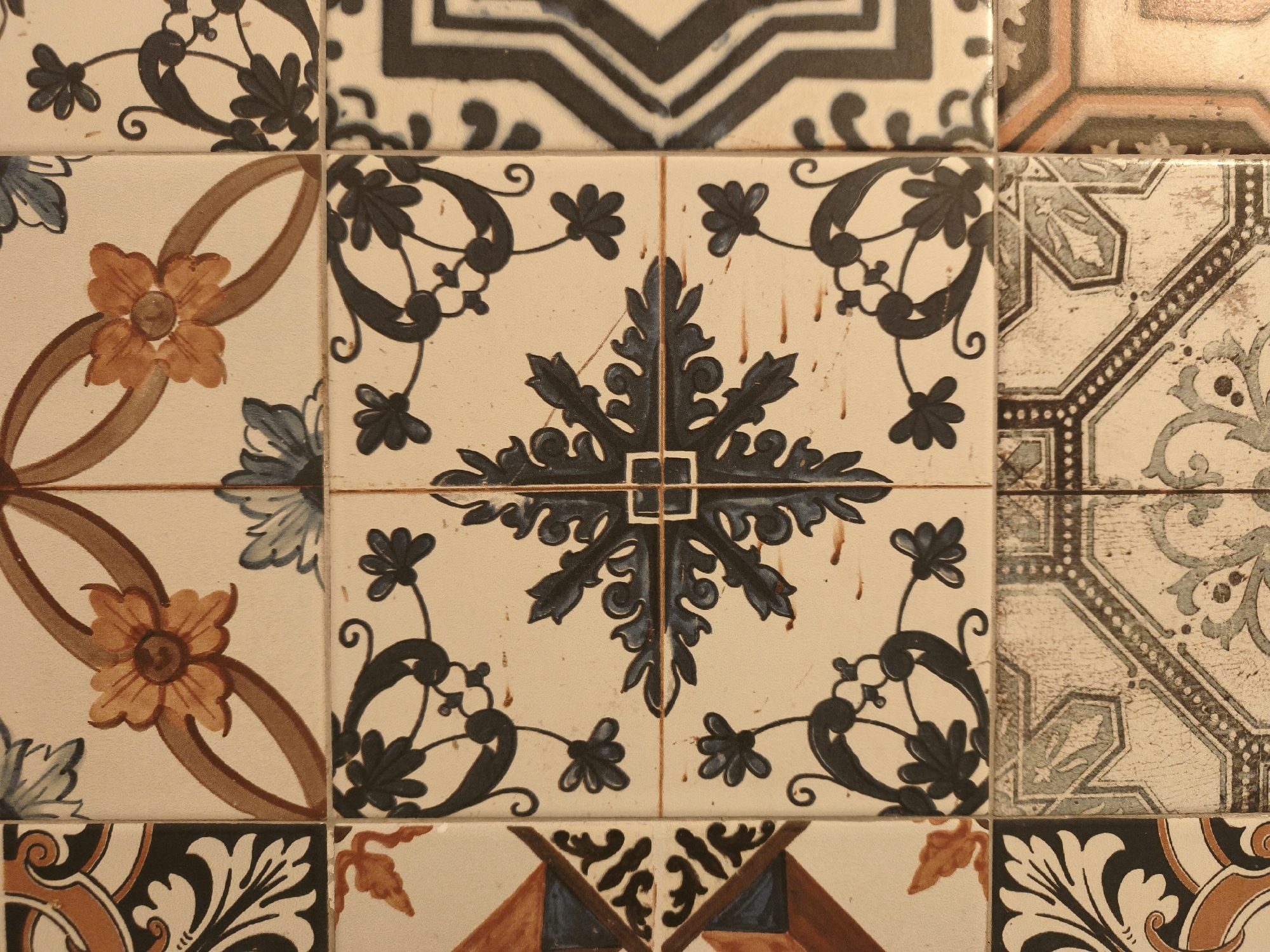 The image shows a close-up of a wall covered in decorative tiles. The tiles feature a variety of patterns, including floral motifs, geometric designs, and stylized foliage. The color palette includes cream, brown, blue, and black. There is no information to suggest a location in the city of Sofia, Bulgaria.