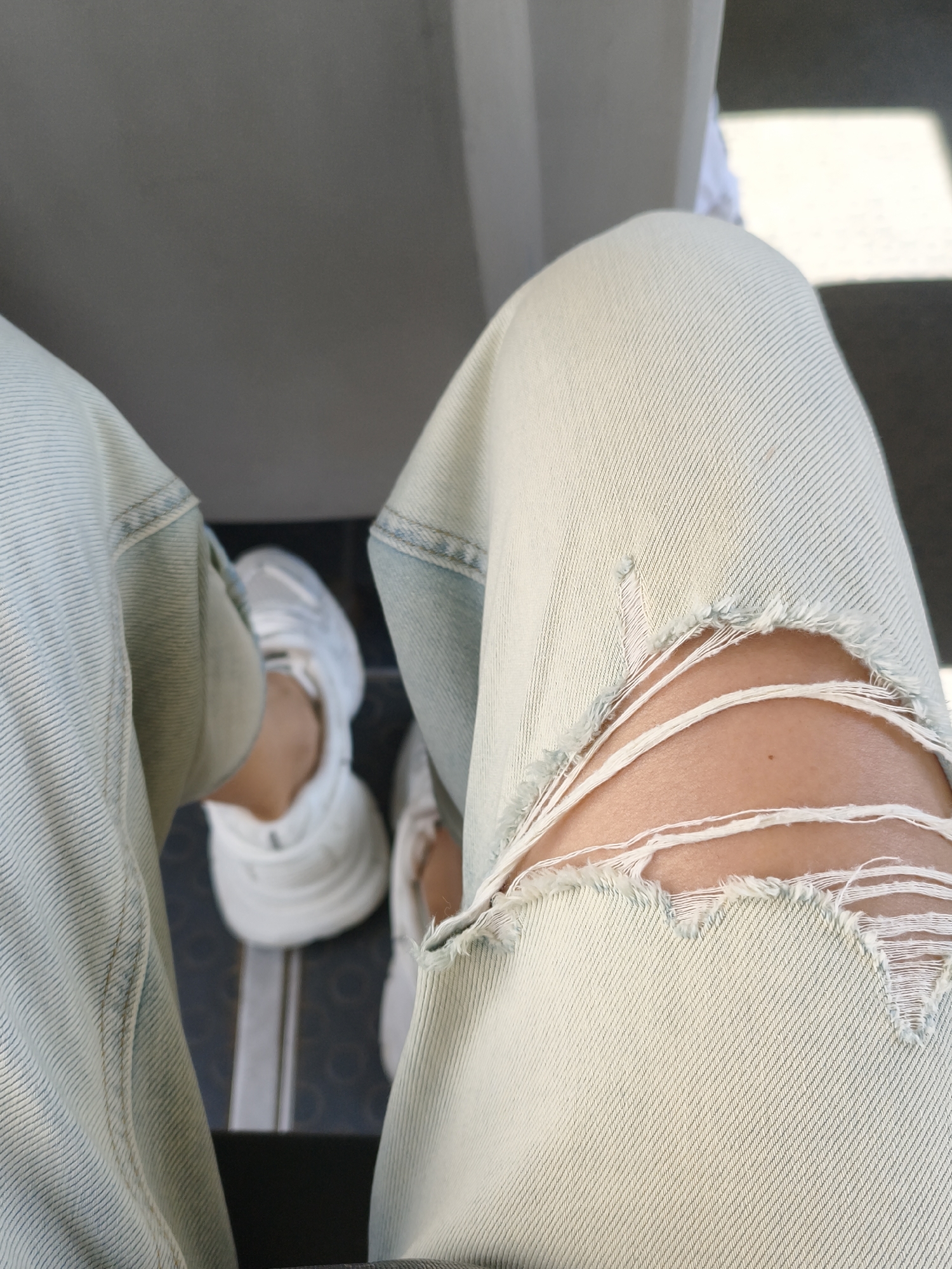 The image shows a close-up of a person's legs and feet, wearing light-colored ripped jeans and white sneakers. The jeans are distressed with large rips on the right knee, exposing skin. The fabric of the jeans has a visible texture and a slightly faded, light blue wash. The sneakers are chunky and appear to be made of a smooth material. The background is out of focus, but suggests an indoor setting, possibly public transport, with a gray seat or wall and a hint of sunlight casting a square shadow on the floor. The floor appears to be tiled with a dark pattern. The perspective is from above, looking down at the person's legs. The overall impression is casual and contemporary. The time of day is likely daytime, given the presence of sunlight. There is no visible text in the image.