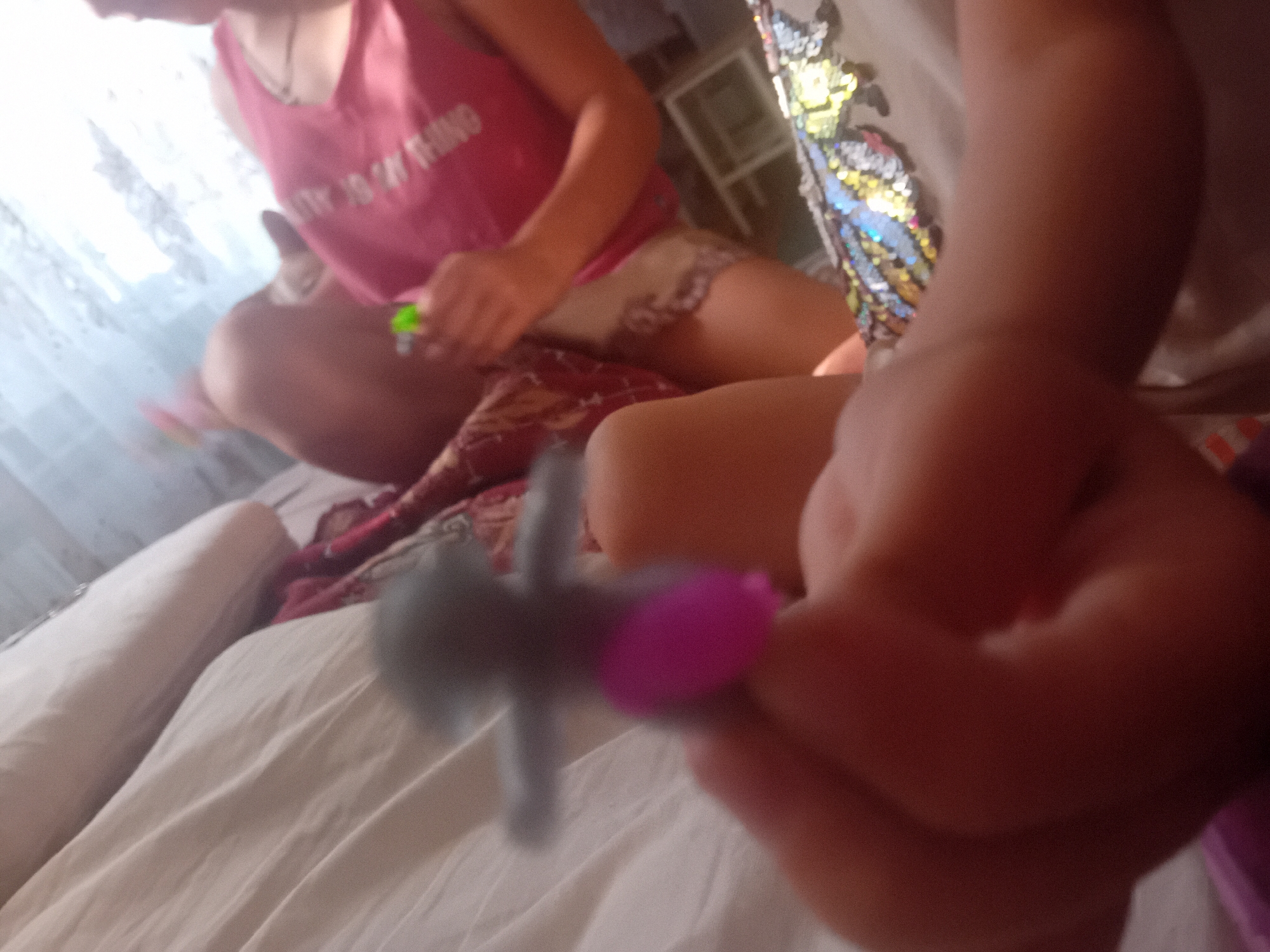 A child's hand is holding a grey and purple fidget spinner in the foreground of the image. The child is wearing a pink tank top with white lettering. In the background, another child is visible, also sitting on a bed. The environment appears to be a bedroom, with white sheets and pillows on the bed. The image does not contain any information to determine the location in the city of Chuhuiv, Ukraine.